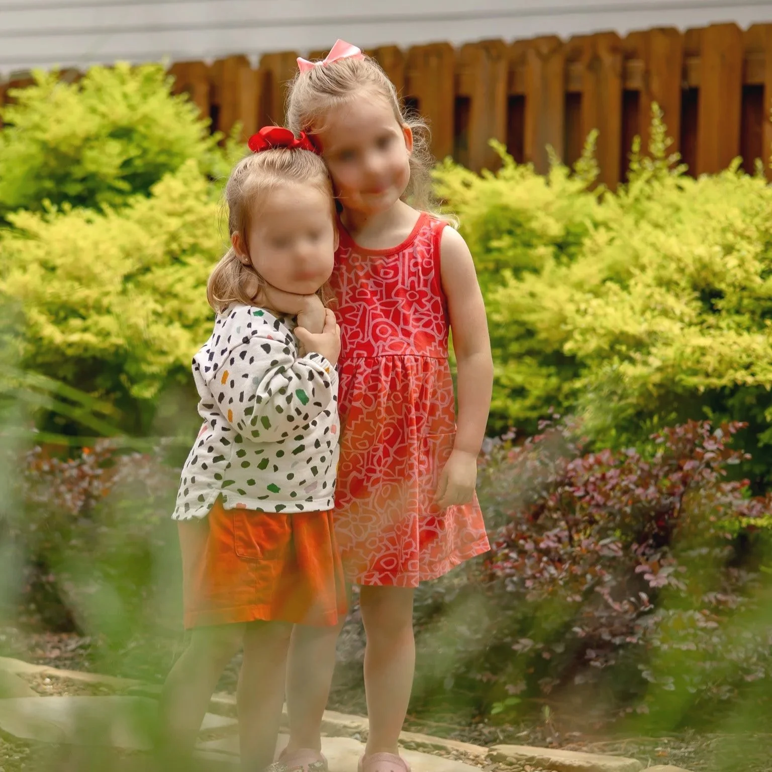 Two young girls standing outdoors in a garden, one with blonde hair in a bun with a pink bow, wearing a red patterned dress, and the other with light brown hair, wearing a white and black polka-dot top with orange shorts.