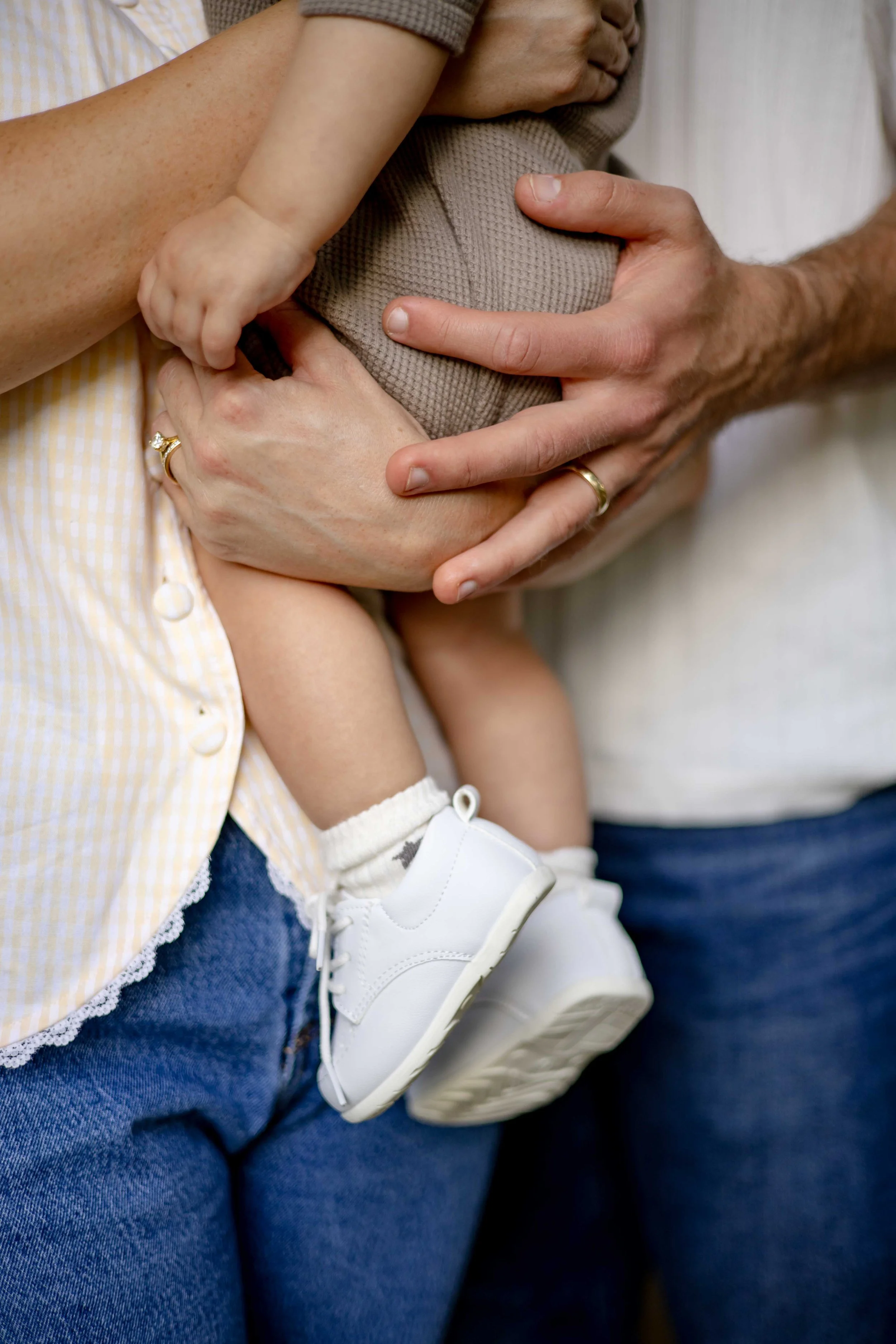 Close-up of a baby with a white shoe, held in a person's arms, with the person's hand on the baby's leg and another person’s hand on the person’s chest, all wearing casual clothing.