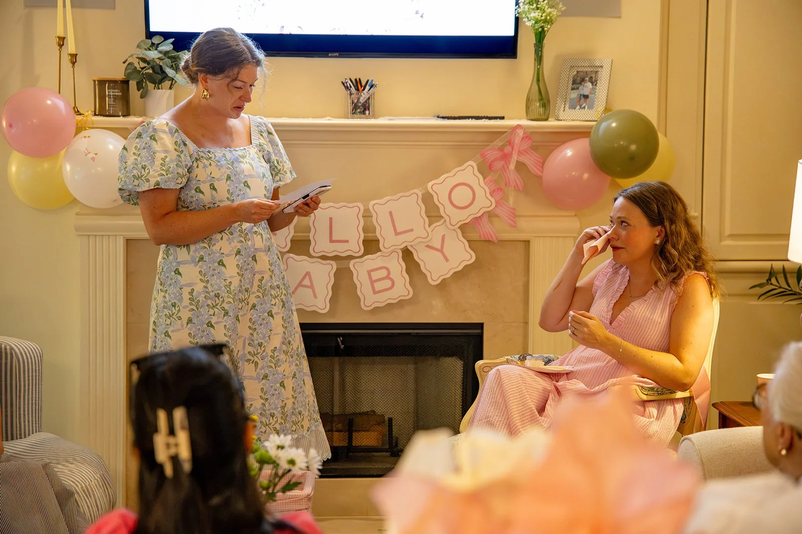 A woman in a floral dress is standing and reading a card at a children's birthday party. A woman in a pink dress is sitting and wiping tears, holding a tissue, near a decorated fireplace with balloons and a pink and white banner.