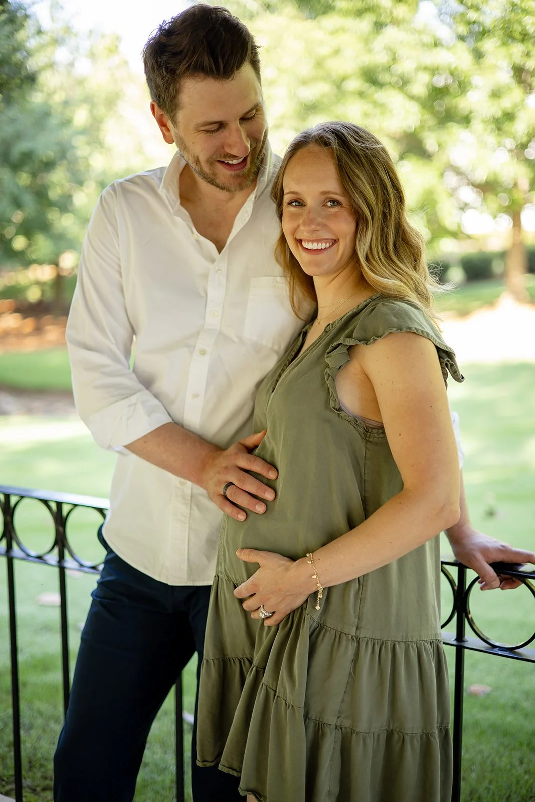 A happy couple standing outdoors, with the man gently touching the pregnant woman's belly. The woman is smiling and wearing a green dress, while the man is in a white shirt. They are near a black metal fence in a park-like setting with green trees and sunlight in the background.