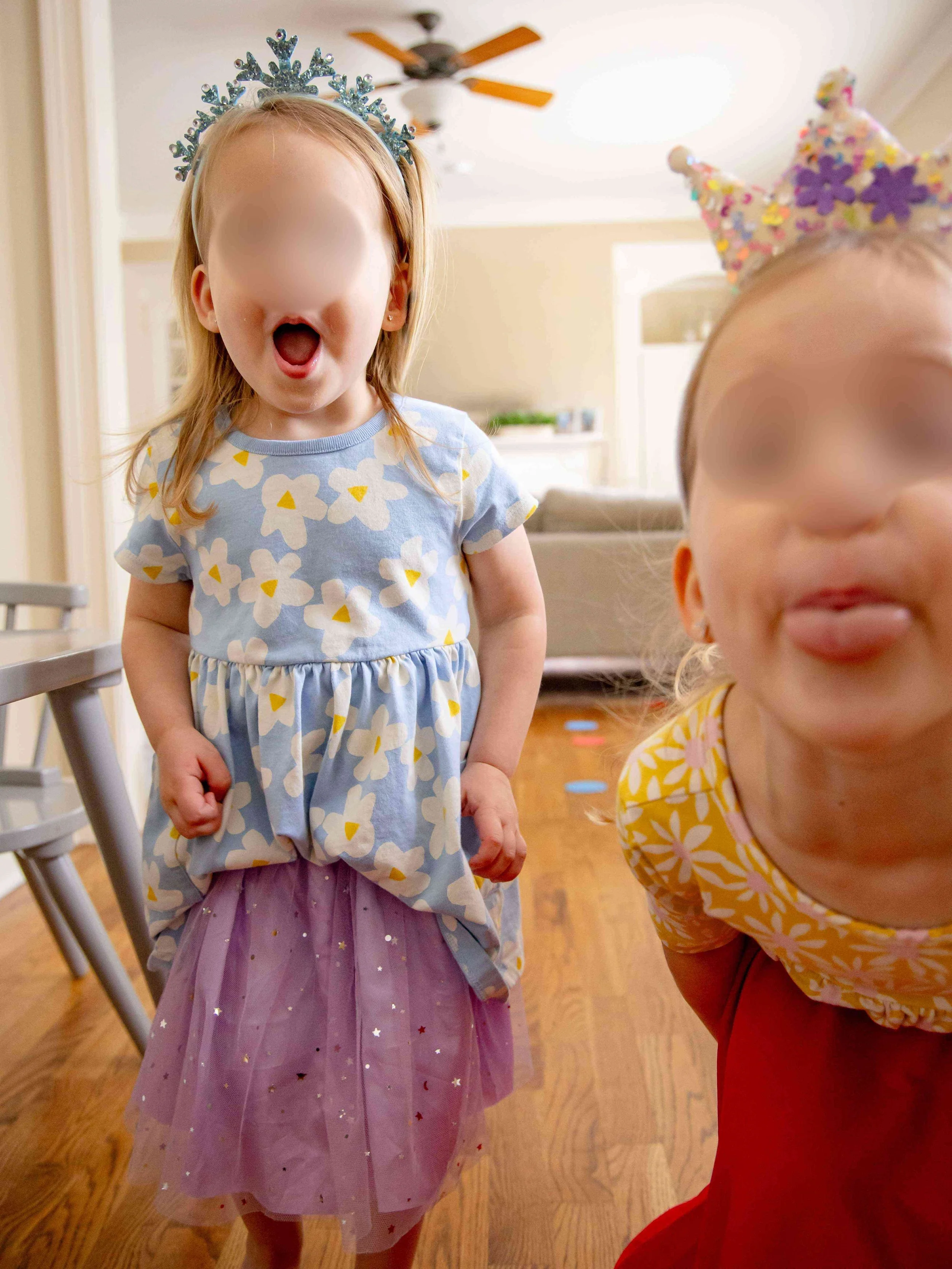 Two young girls wearing birthday crowns and celebrating, one in a floral dress with a purple tulle skirt, and the other in a yellow and orange floral top with red pants, indoors in a room with hardwood flooring.