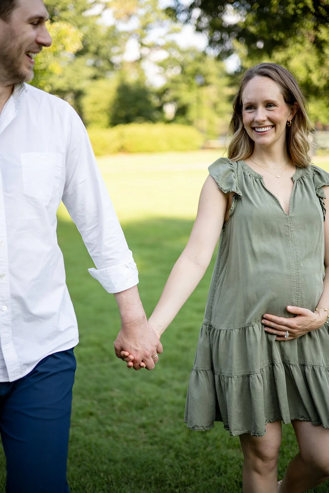 A pregnant woman holding her belly and smiling, holding hands with a man outdoors in a park, with trees and grass in the background.