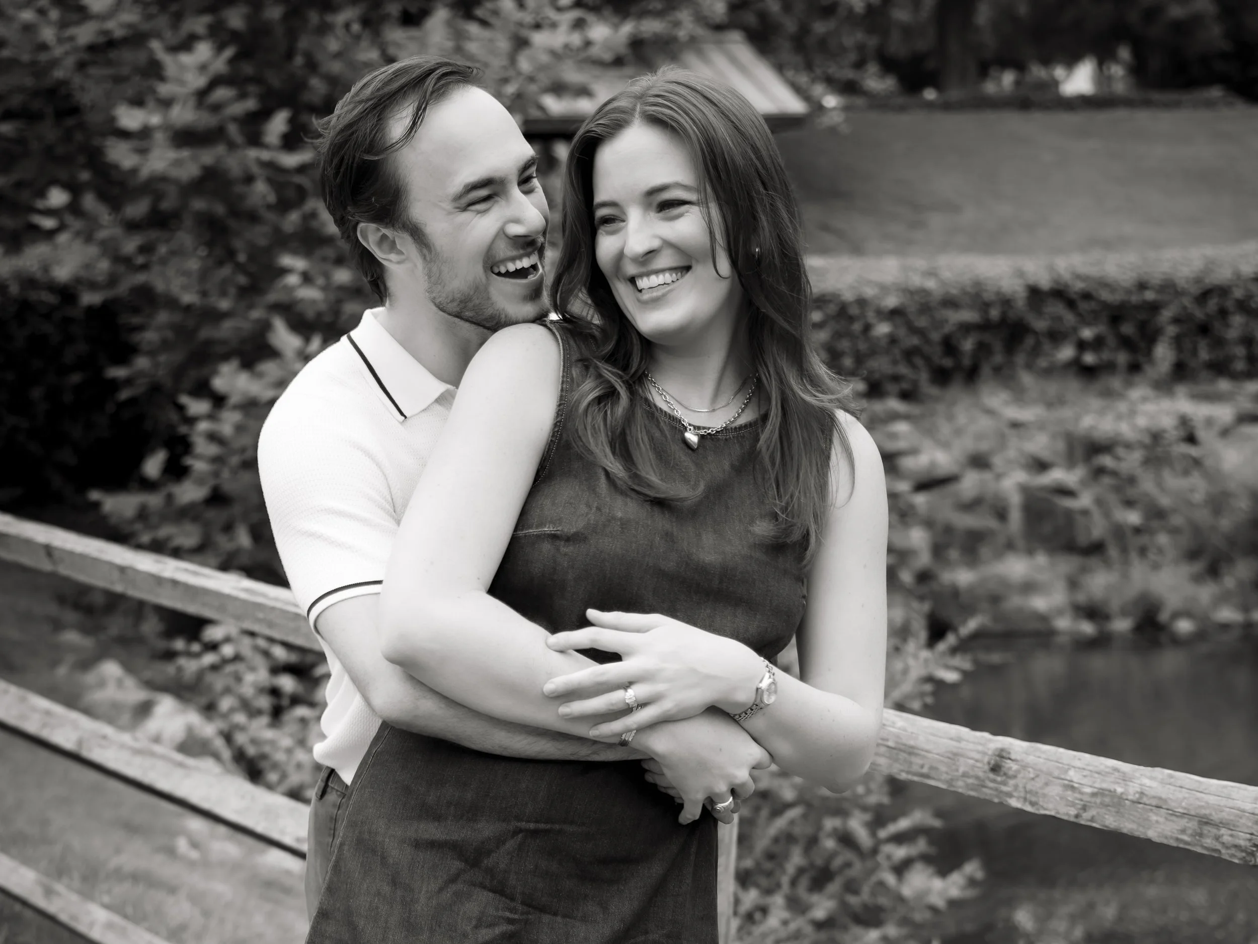 A happy young couple embraces in a park, smiling and looking at each other, surrounded by trees and foliage.