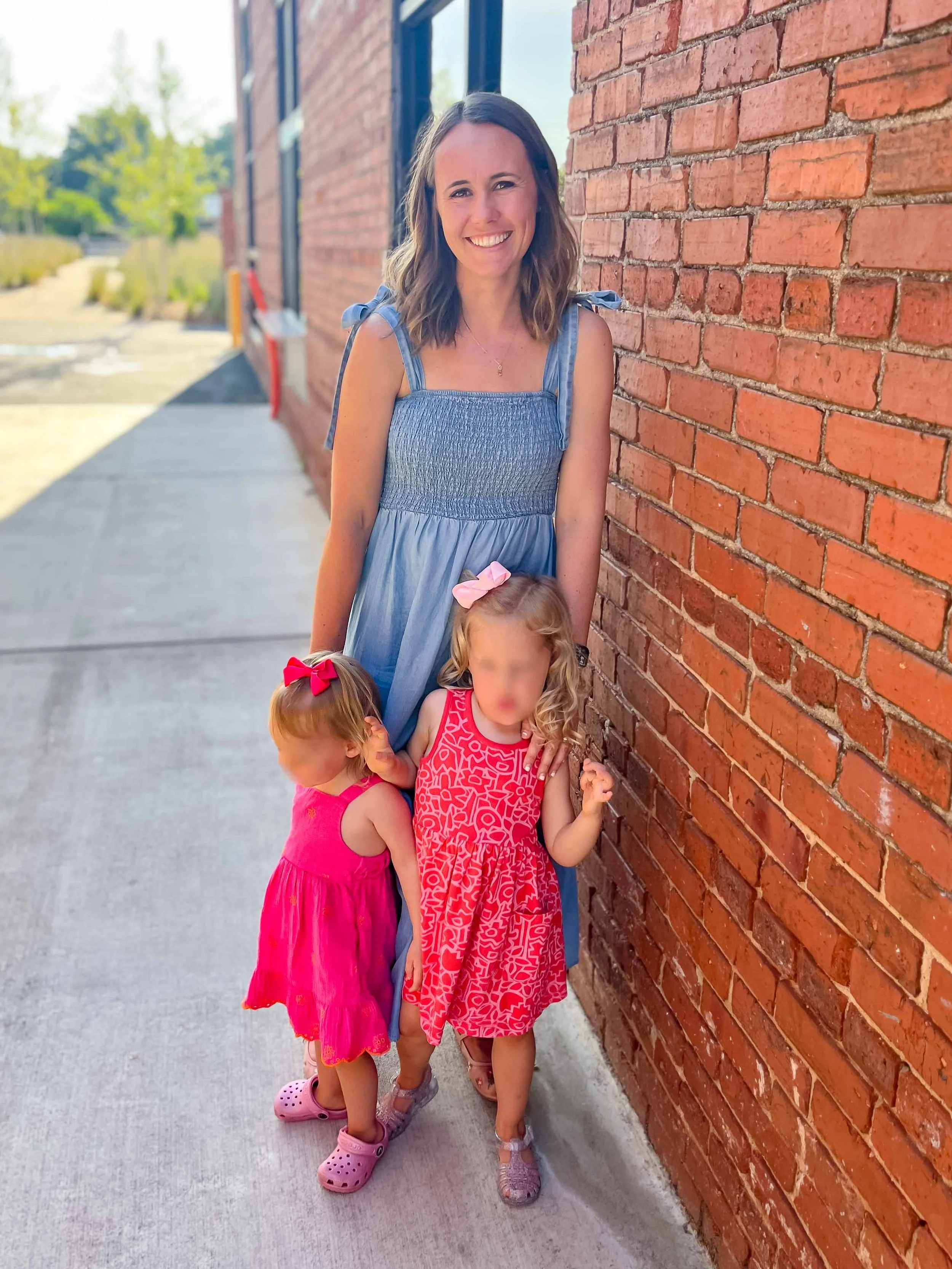 A woman smiling standing next to a brick wall with two young girls, one in a pink dress with a red bow and the other in a red and white patterned dress with a pink bow, on a sidewalk outdoors on a sunny day.
