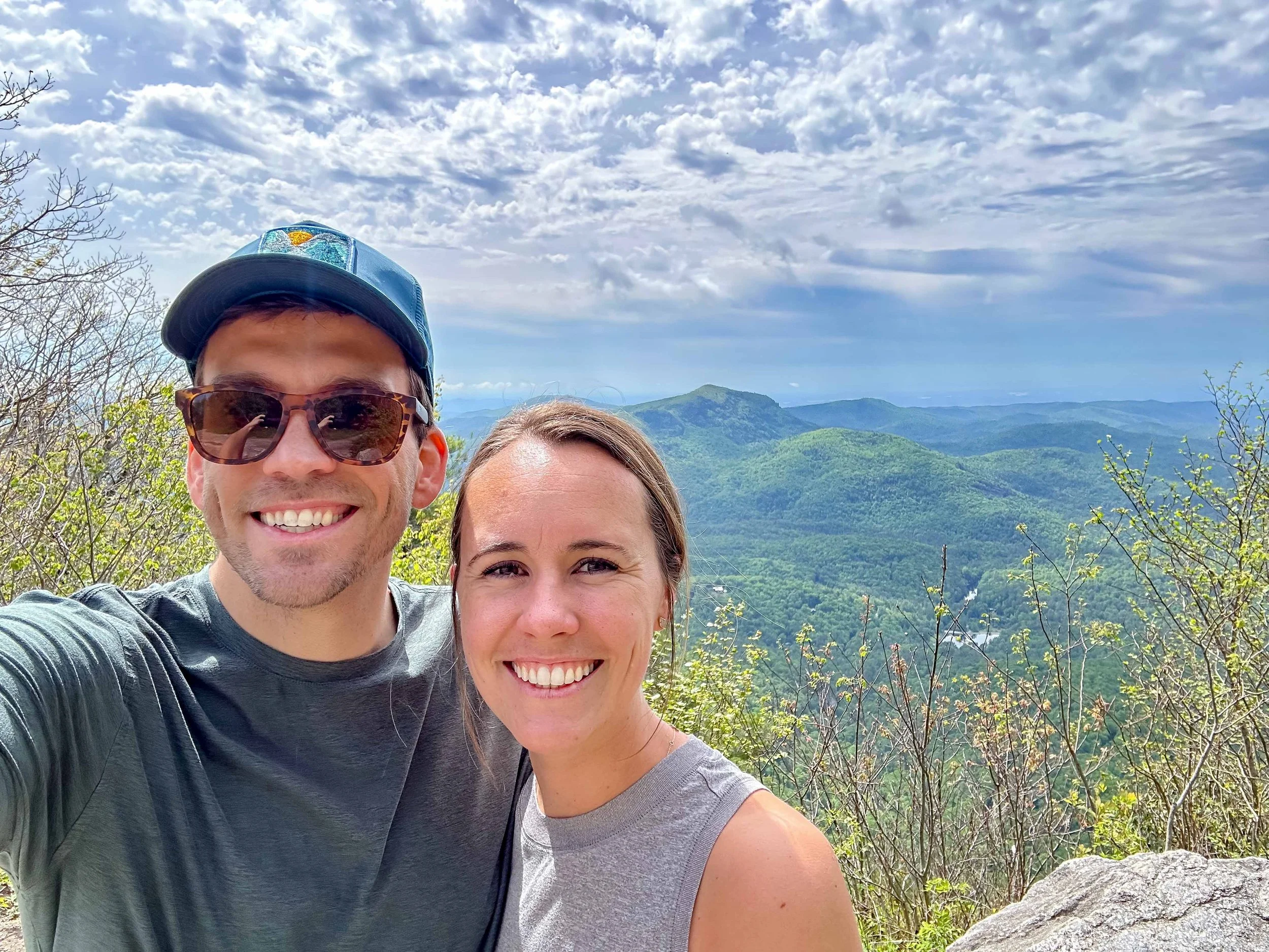 A smiling man wearing sunglasses and a blue cap, and a smiling woman, taking a selfie together outdoors with a mountain landscape, cloudy sky, and trees in the background.