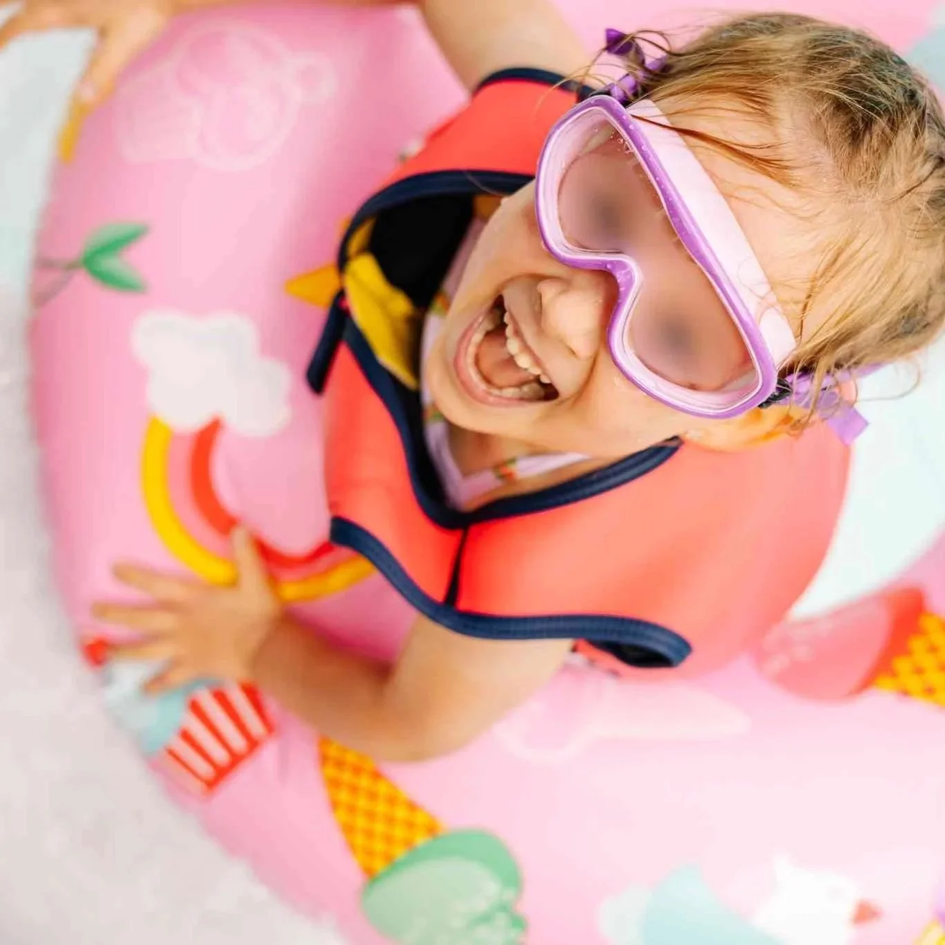 A young girl with wet hair wearing pink goggles, a colorful life jacket, and a patterned swimsuit, smiling and playing in a pink float ring decorated with colorful elements.