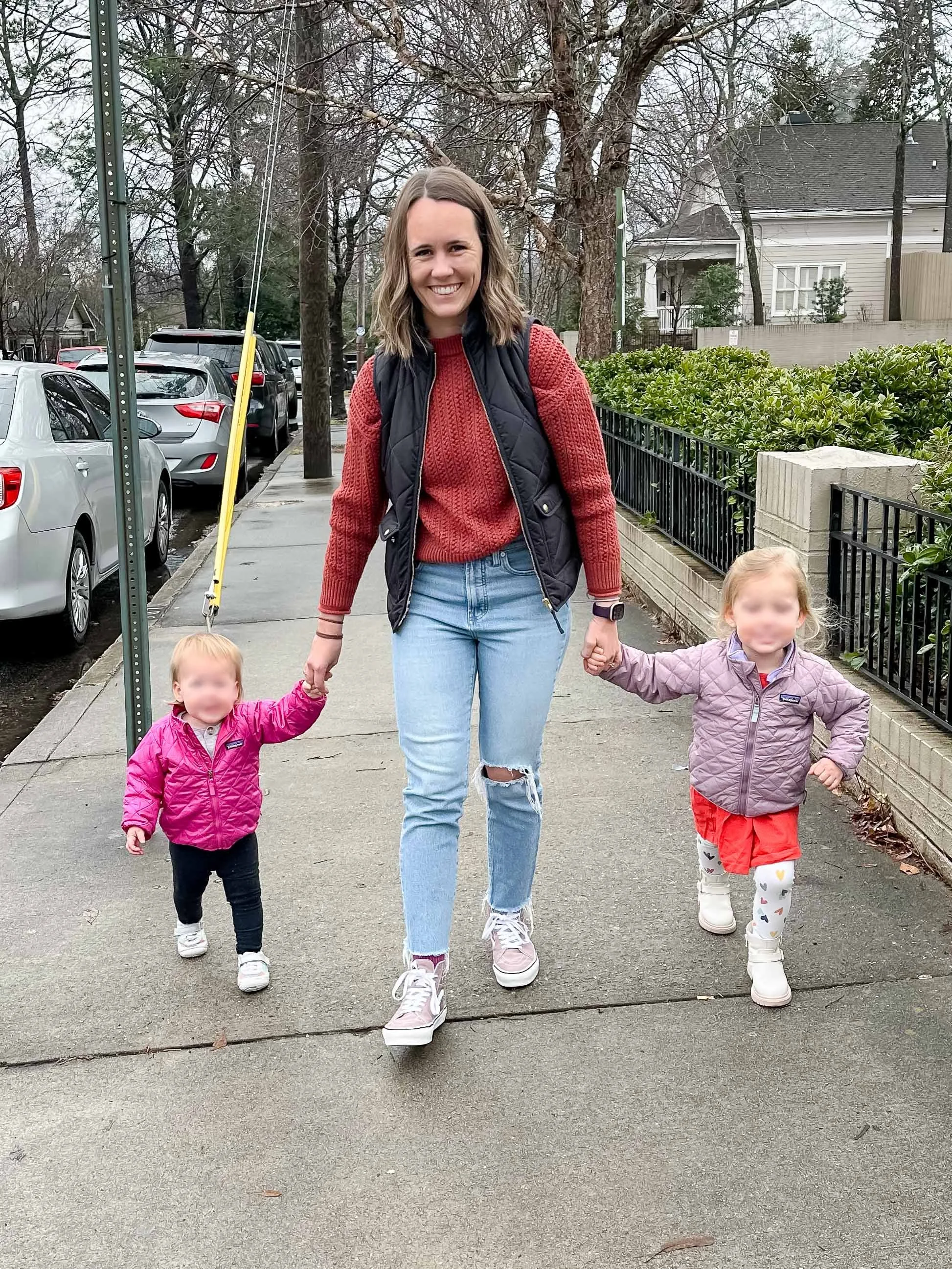 A woman walking with two young girls on a sidewalk. The woman is wearing a red sweater, a black vest, and ripped jeans. The girl on the left is wearing a pink jacket and black pants, and the girl on the right is wearing a purple jacket and white pants with colorful hearts. It is daytime, and there are parked cars, trees, and houses in the background.