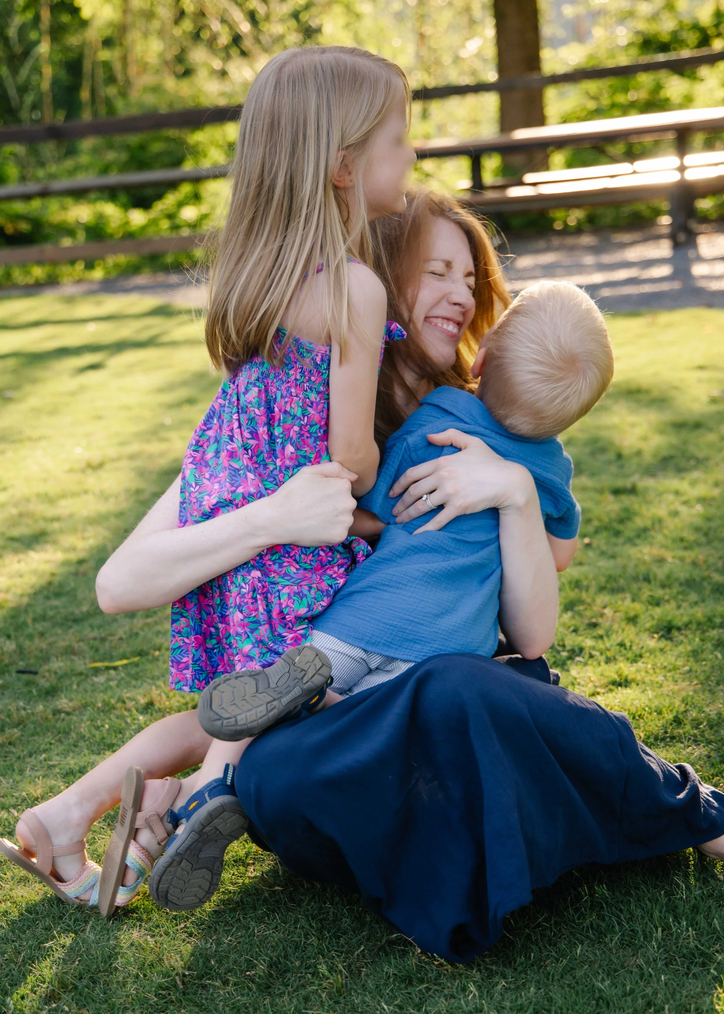 A woman sitting on grass outdoors, laughing while holding a young boy, with a girl standing on her lap, all smiling and enjoying a sunny day in a park.