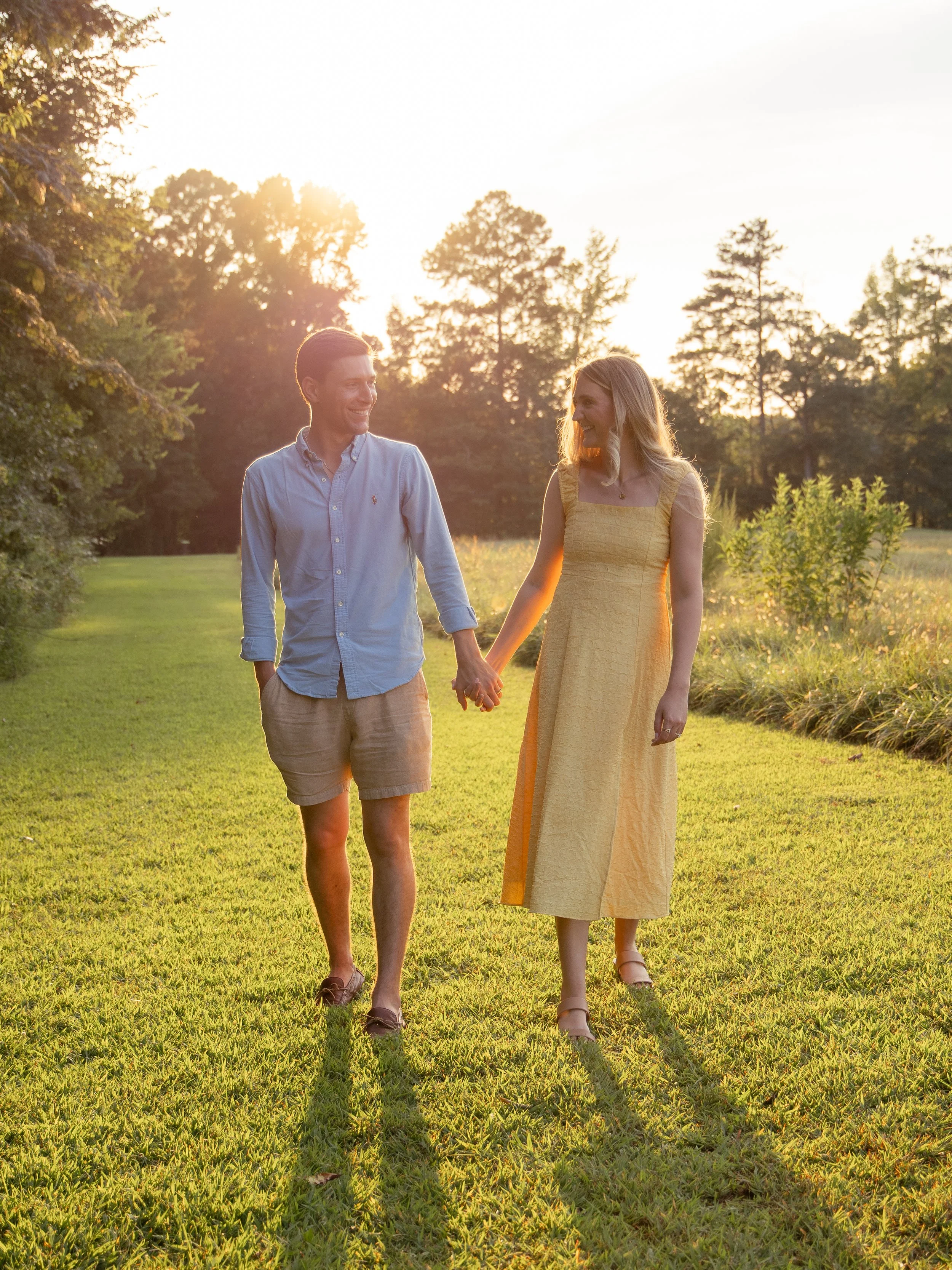 A man and woman holding hands and walking through a sunlit park during sunset, surrounded by green grass and trees.
