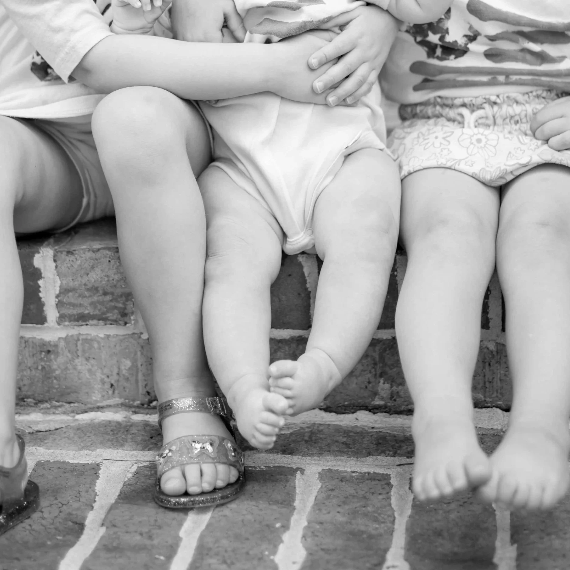 Close-up black-and-white photo of children sitting on brick stairs. Focus on their legs and feet, with some children wearing shiny sandals. One child's arm and hand are visible, resting on their lap.