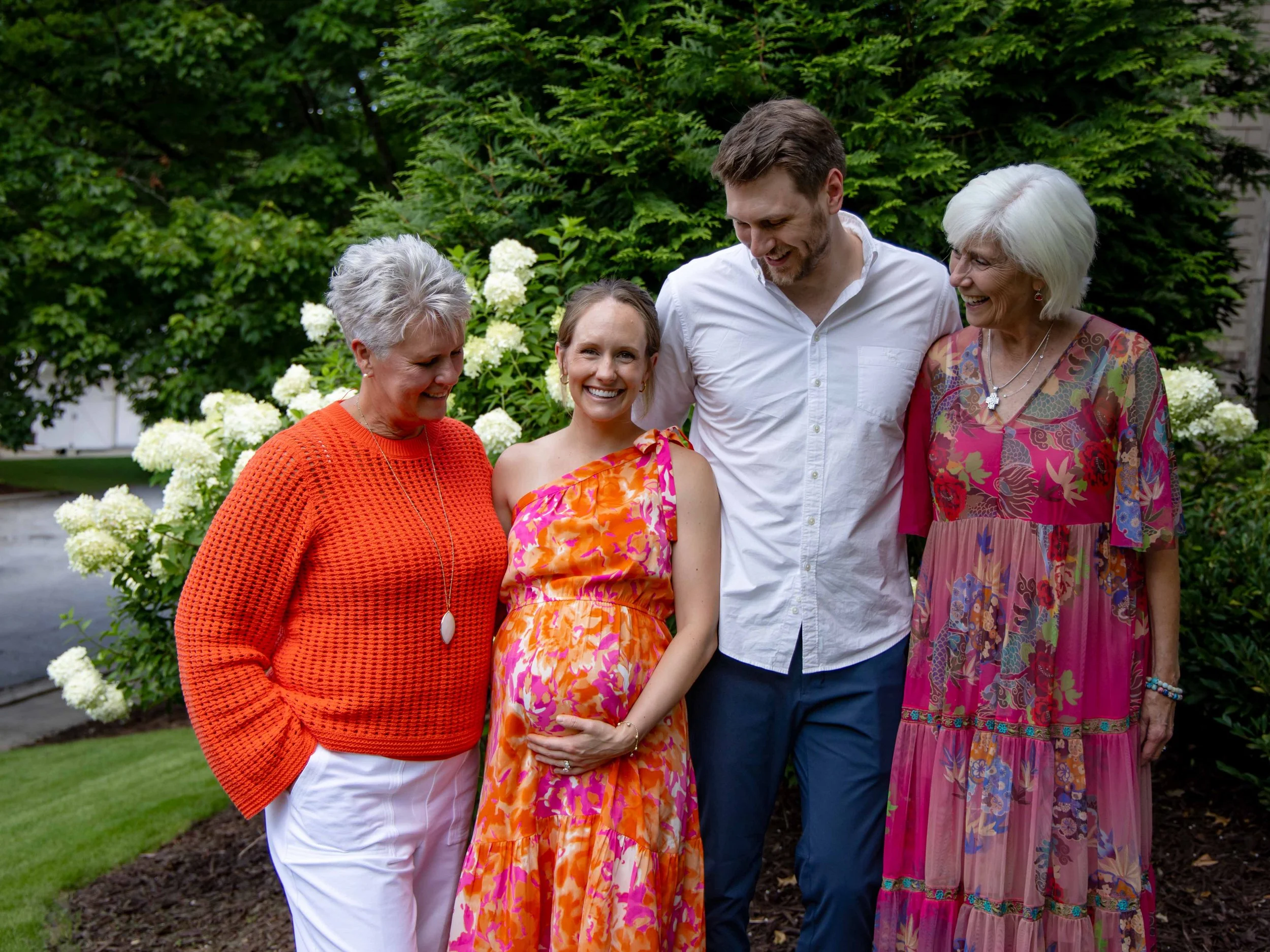 A group of five people, including a pregnant woman, smiling and standing closely together outdoors among green trees and white flowering bushes.