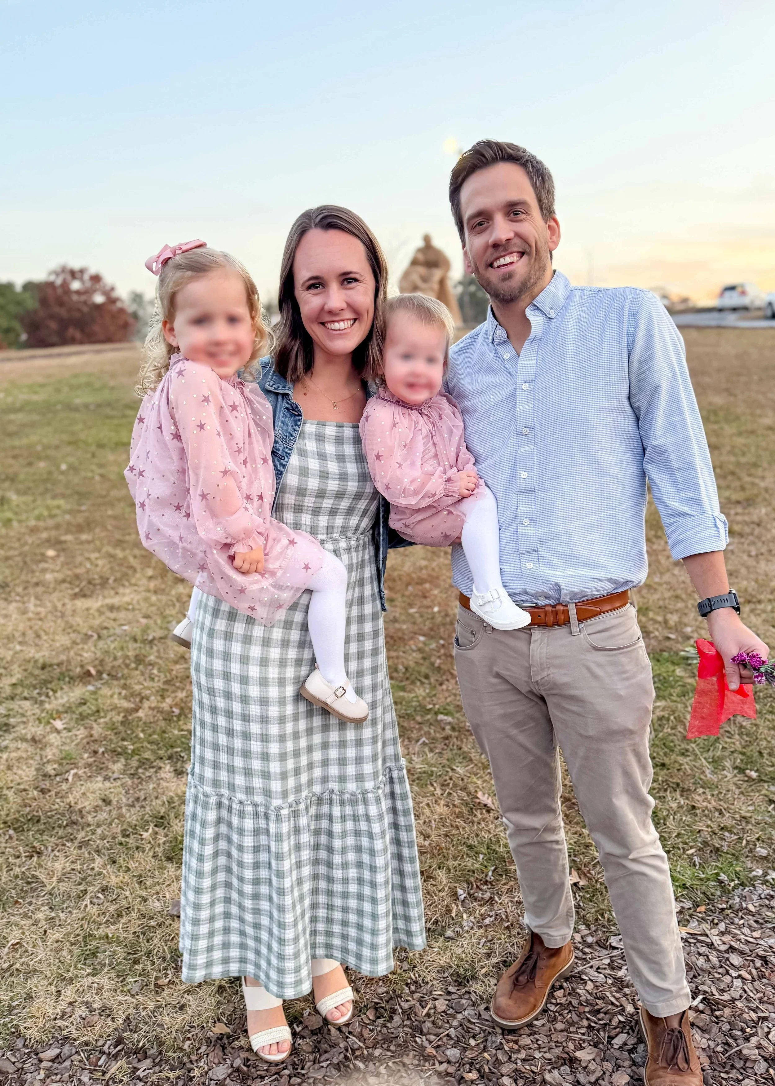 A smiling family of four outdoors at sunset, with a father, mother, and two young daughters, holding flowers and dressed casually.