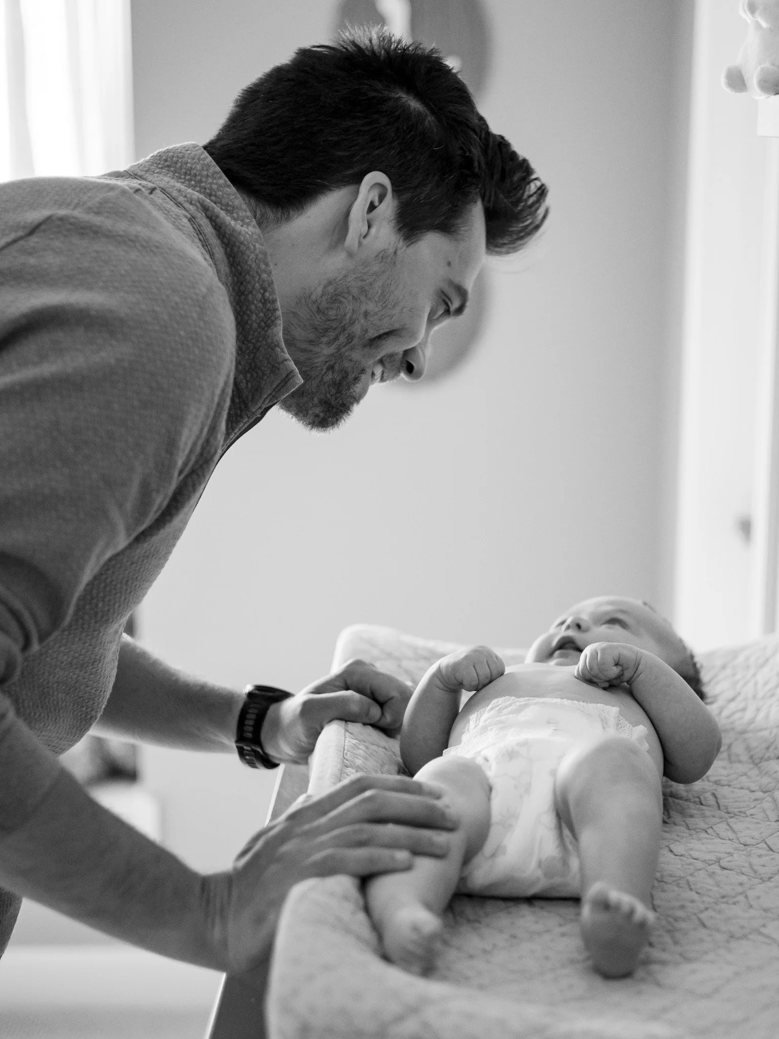 A man with dark hair and a beard leaning over a baby lying on a changing table. The man is holding the baby's hand and looking at the baby with a smile. The baby is looking up at the man, wearing a diaper.