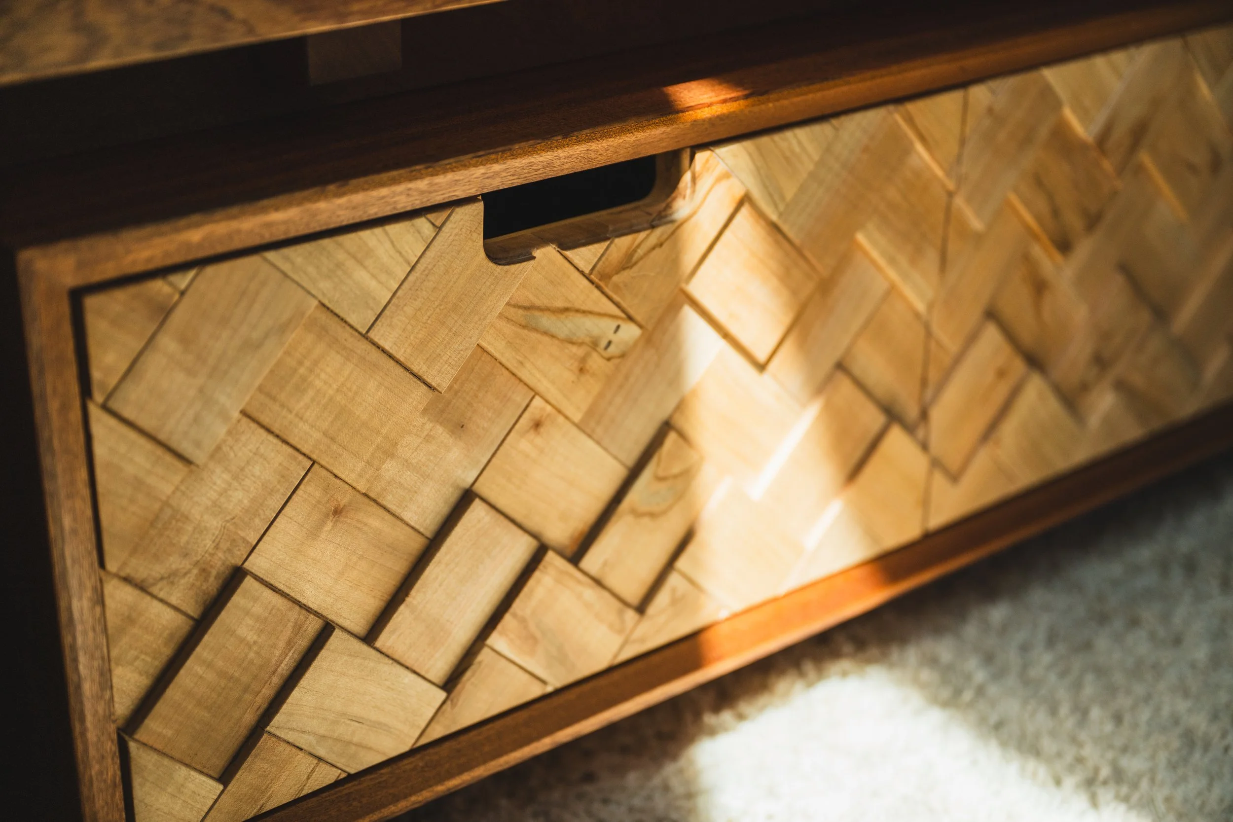 Close-up of a wooden furniture piece with a herringbone pattern and a drawer handle, viewed from an angle.