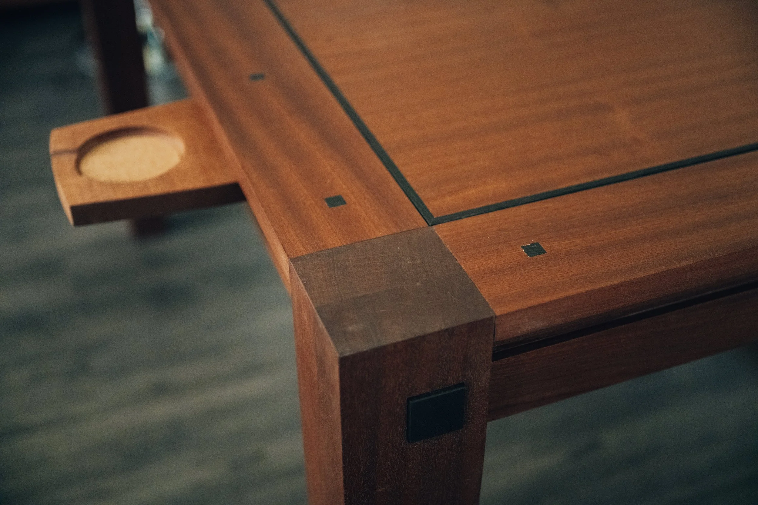 Close-up of a wooden table with a light brown finish, featuring black inlaid lines and small black square accents, with a corner of a wooden chair visible on the left.