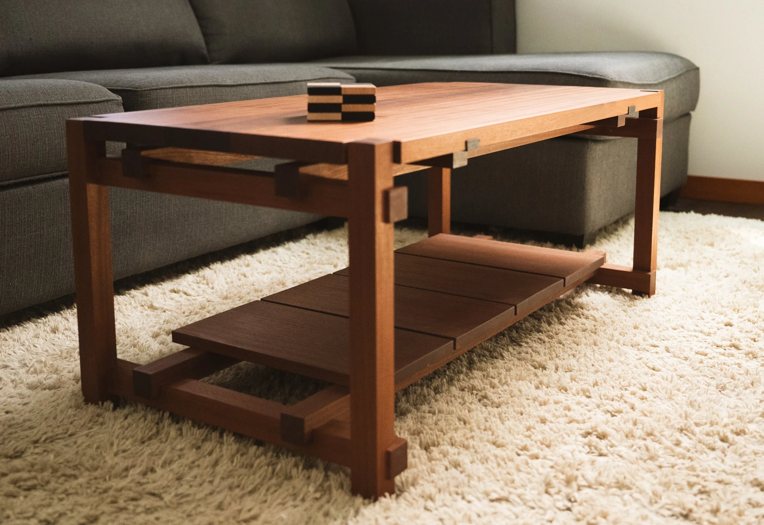 A wooden coffee table with a rack underneath, placed on a cream-colored shaggy rug in front of a dark grey sofa.