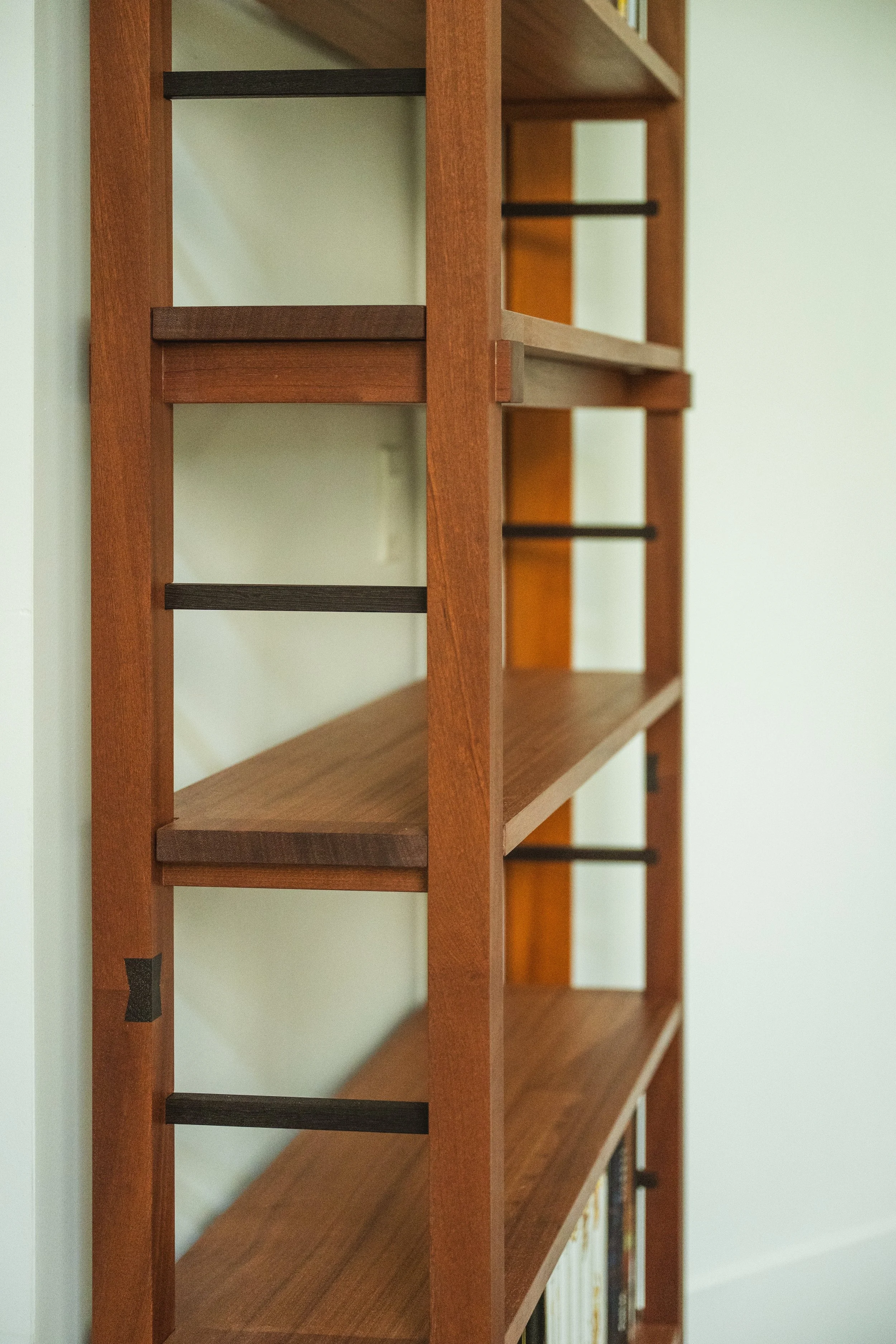A wooden bookshelf with black support bars and a few books on the bottom shelf, positioned against a white wall.