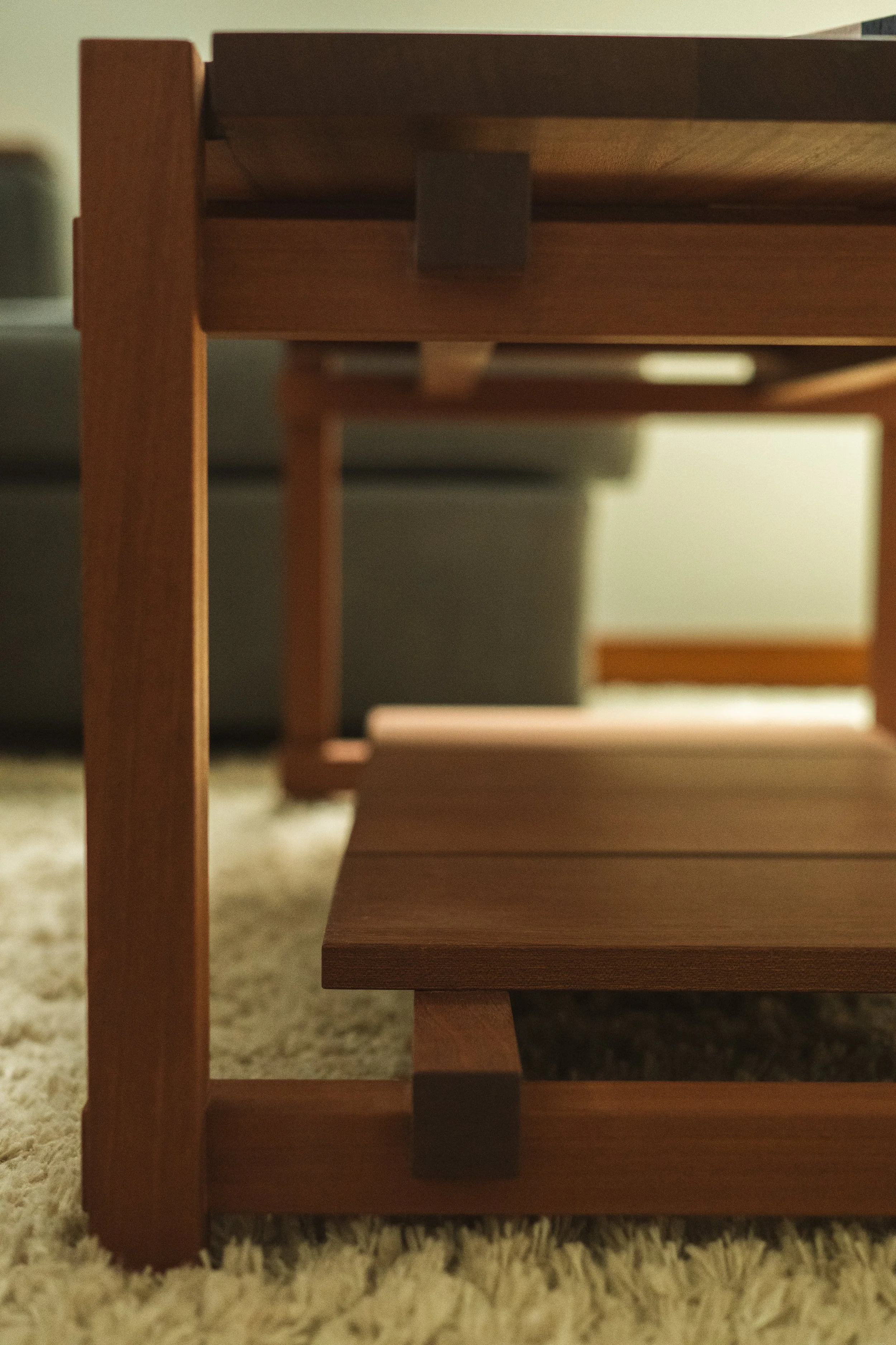 A close-up view of a wooden table's corner, showing the tabletop, legs, and support structure, situated on a beige carpeted floor.