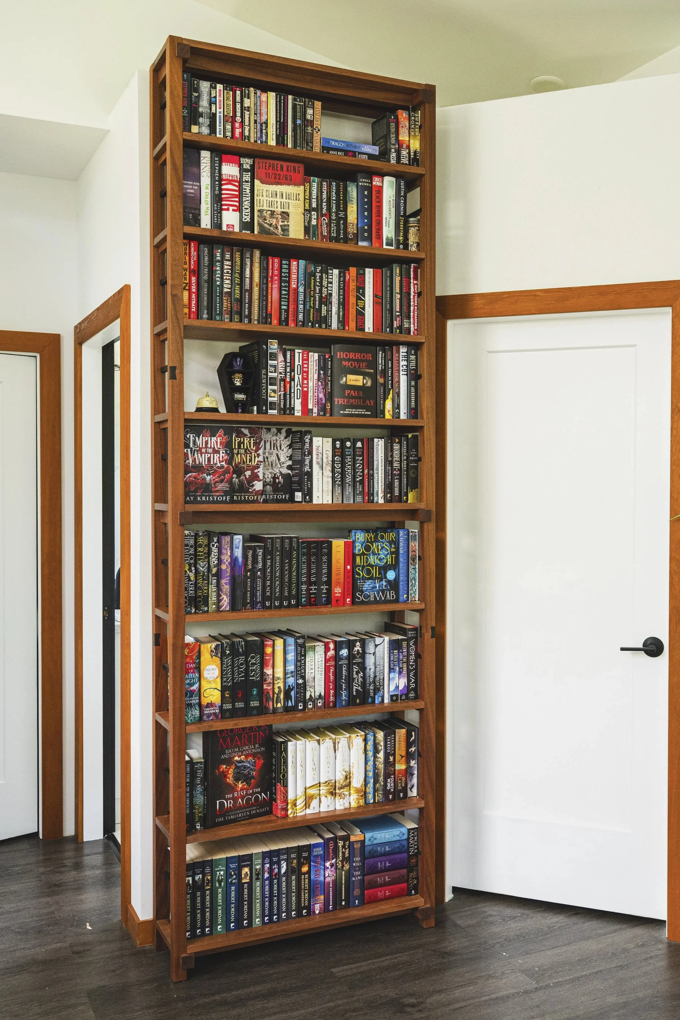 A tall wooden bookshelf filled with books, with a variety of colors and sizes, located in a room with white walls and dark hardwood floors. Part of a white door is visible on the right, and a door with a glass panel is on the left.