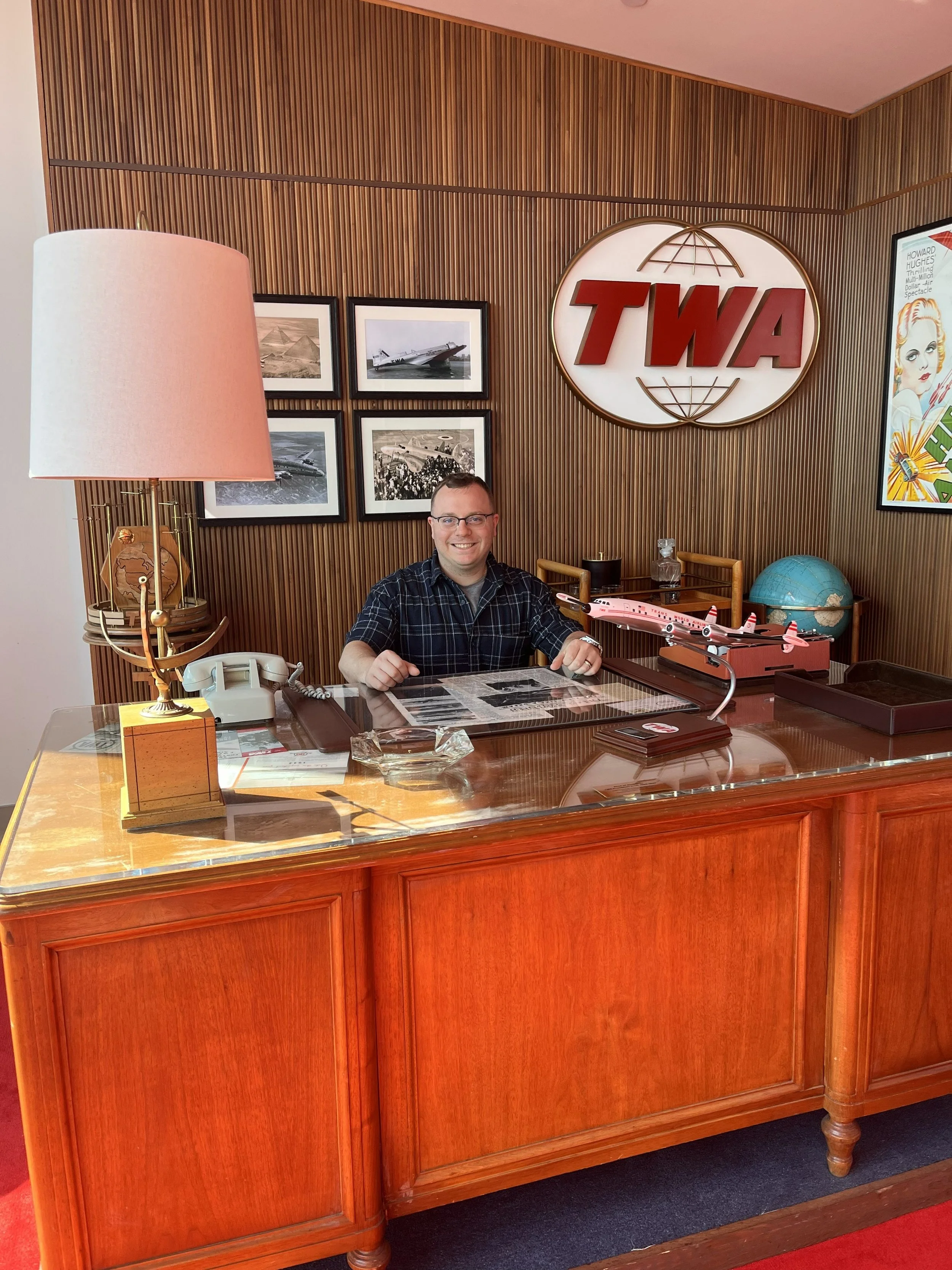 Smiling man sitting behind a wooden reception desk with model airplanes, a vintage telephone, and a globe, in a room with wooden paneled walls and aviation pictures, and a large TWA logo sign on the wall.