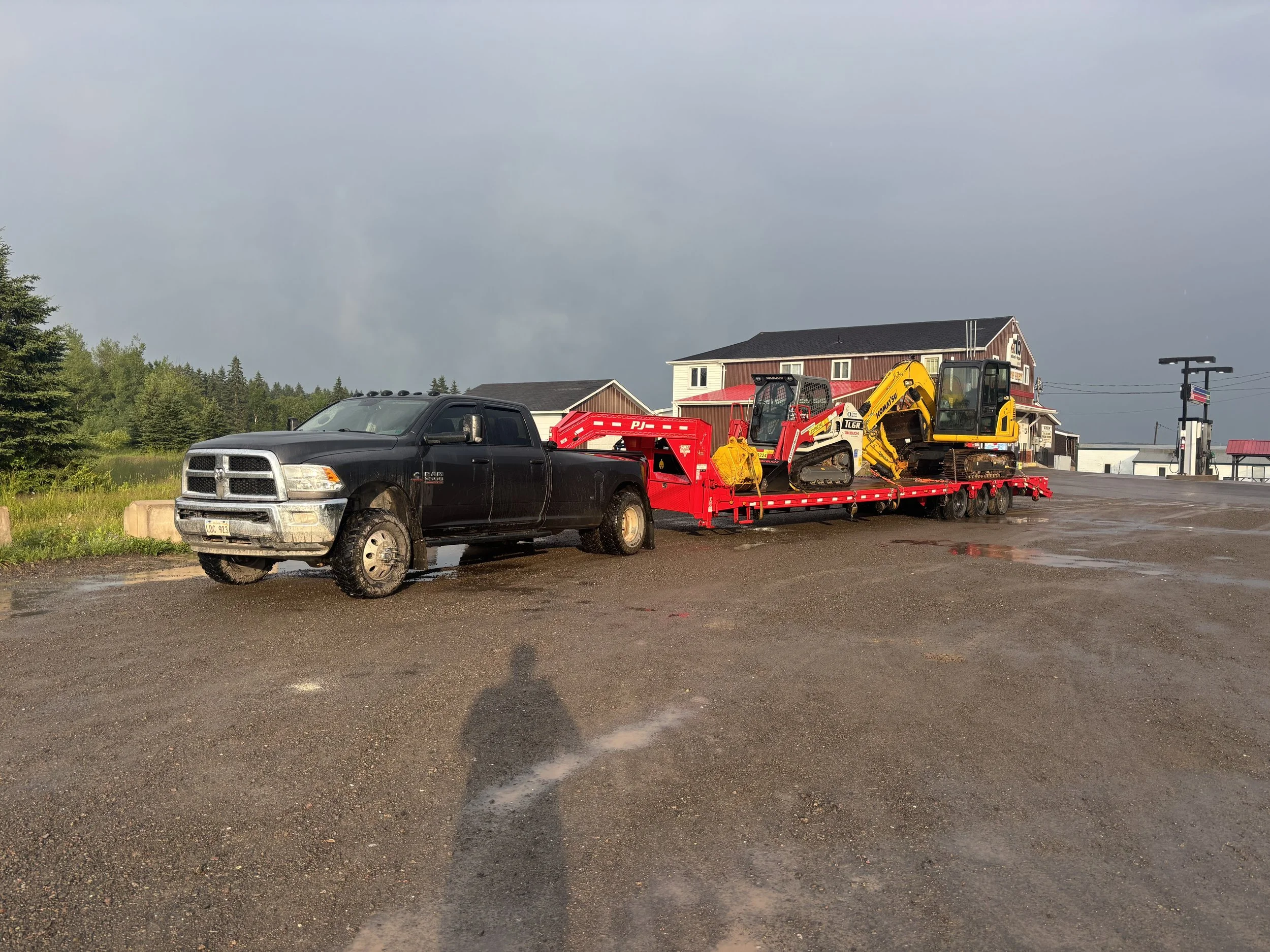 A black pickup truck pulling a red flatbed trailer carrying construction equipment, including a yellow backhoe, parked on a wet gravel lot outside a building under a dark cloudy sky.