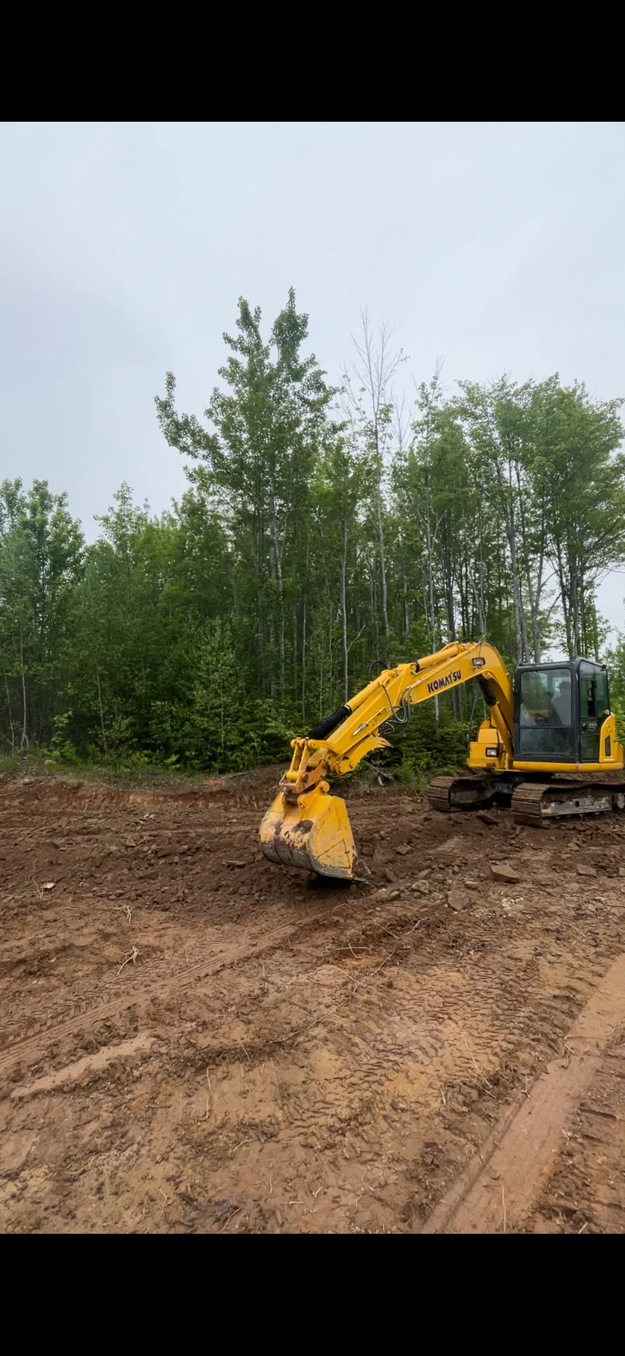 A yellow Komatsu excavator working on a dirt construction site with green trees in the background.