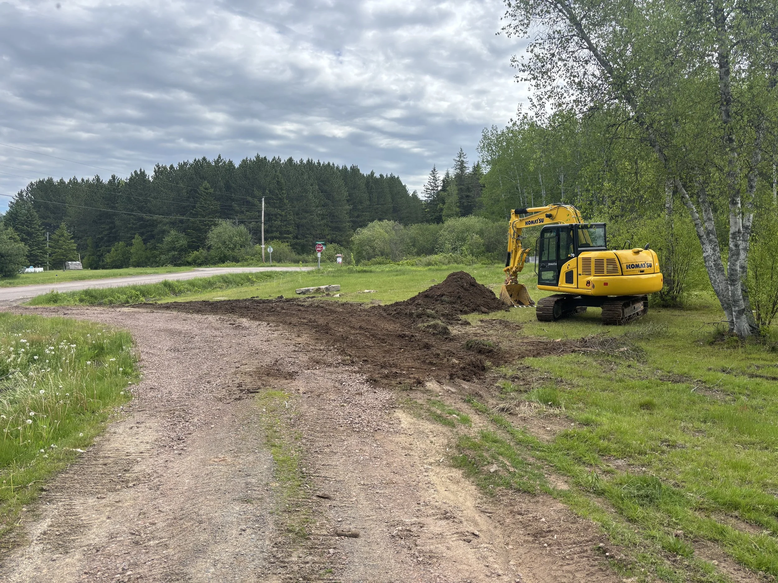 A construction site with a small yellow Komatsu excavator on the right, a pile of dirt in front of it, and a dirt path leading towards a road. Trees with green leaves are in the background under a cloudy sky.