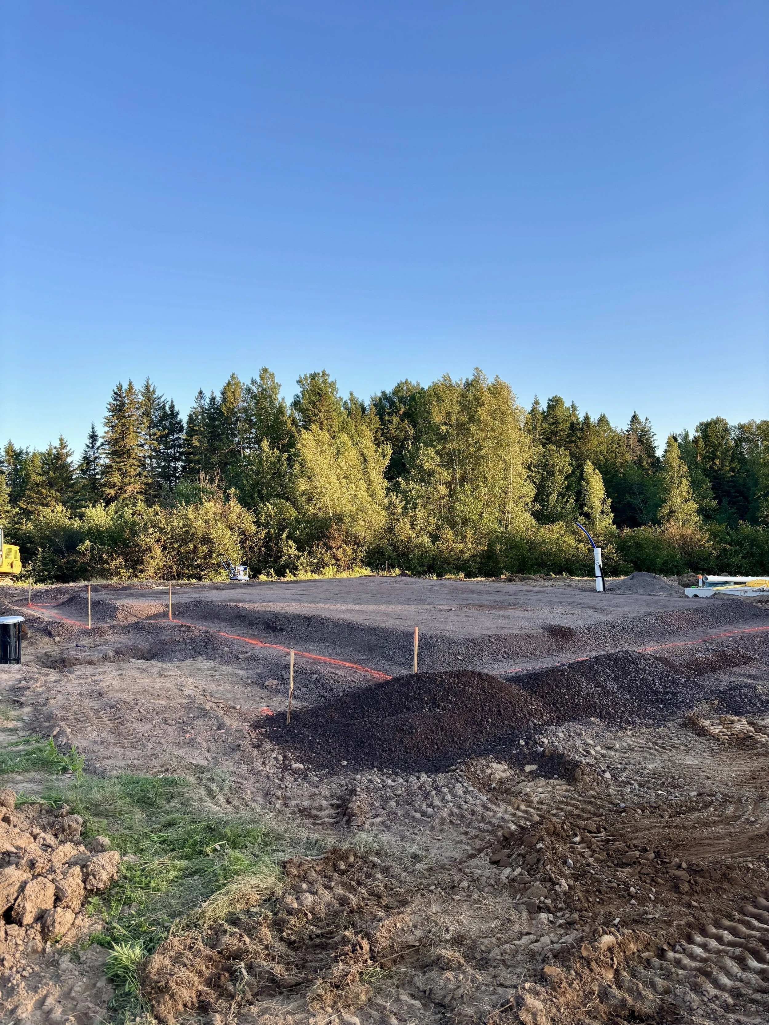 Construction site with cleared land and built pad, construction equipment, and dirt piles, surrounded by a forest with green trees under a clear blue sky.