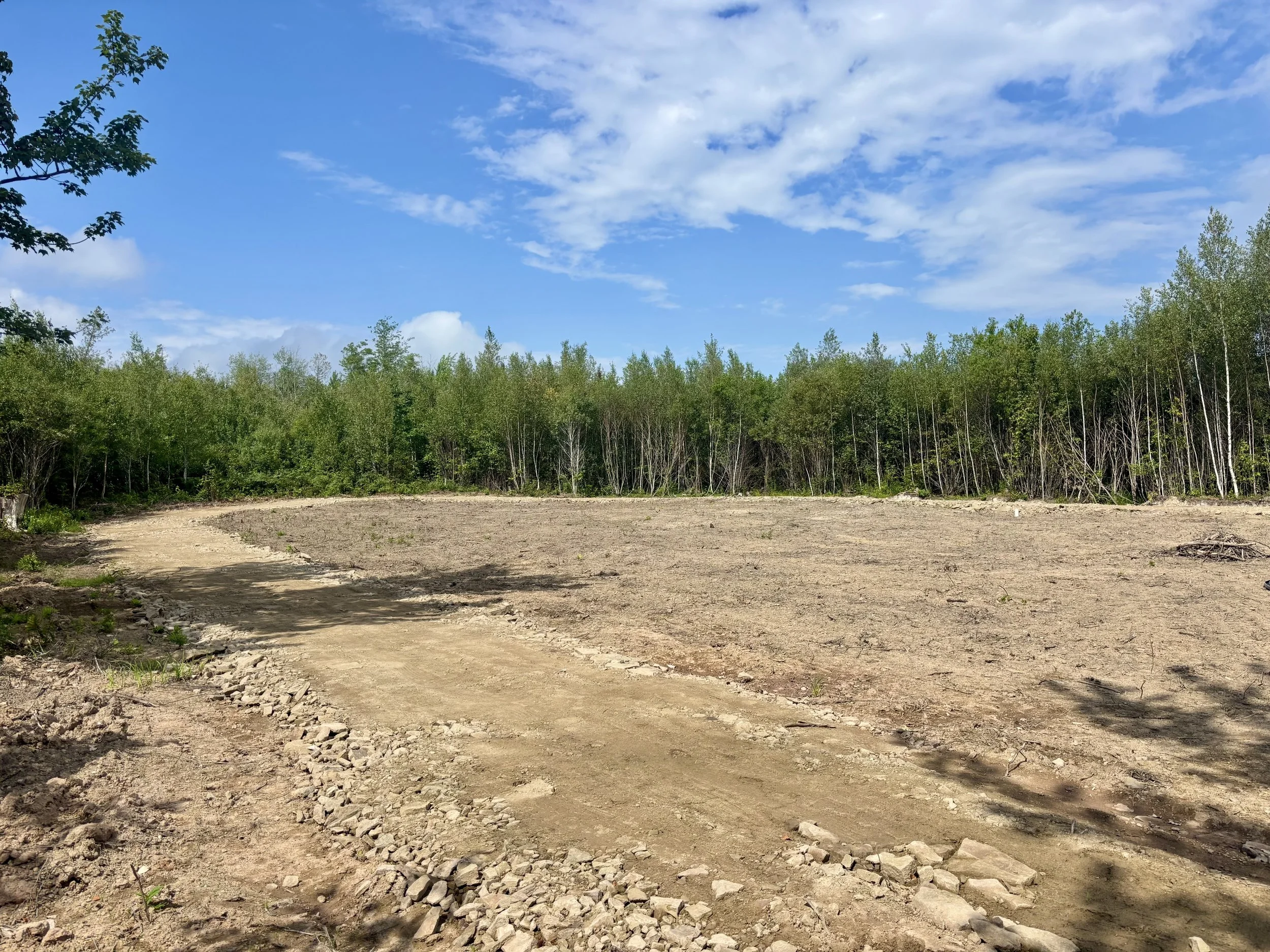 A cleared dirt plot in a rural area with sand stone driveway wrapping a treeline in the background under a partly cloudy blue sky.