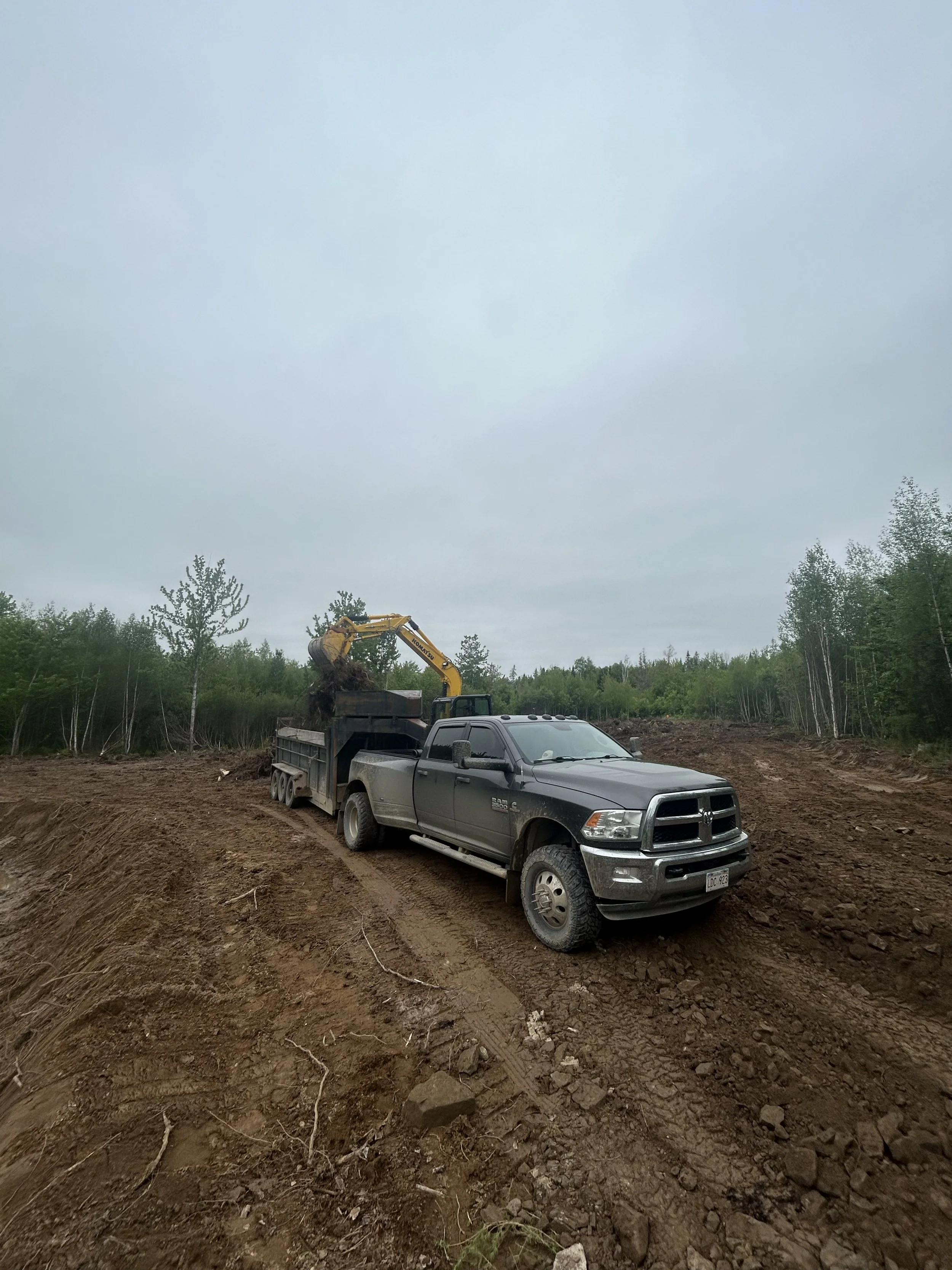 A construction site with a black pickup truck and a yellow excavator digging up dirt under a cloudy sky, surrounded by trees.