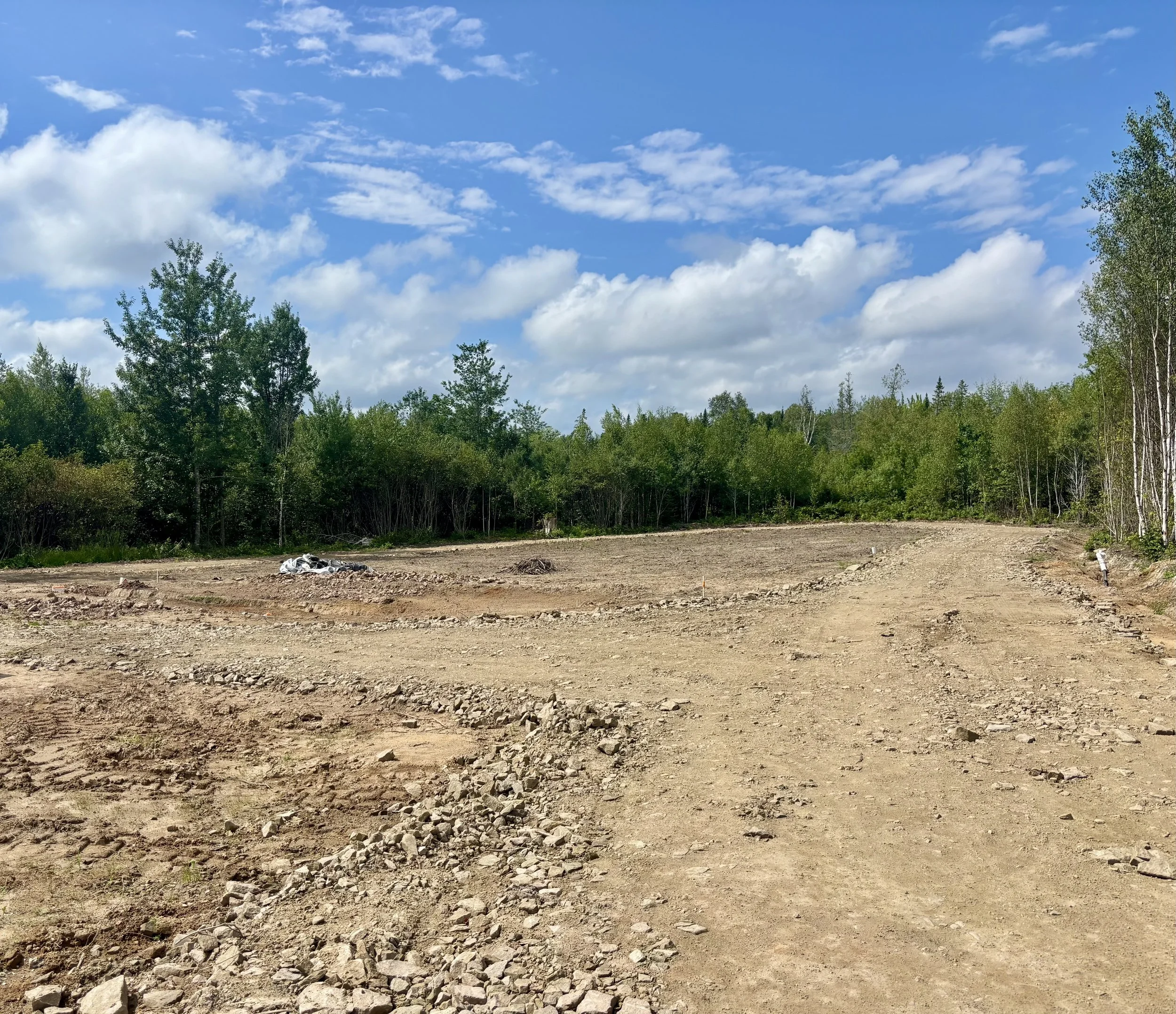 An open construction site with dirt and rocks in the foreground, dense green trees in the background, and a partly cloudy blue sky above.