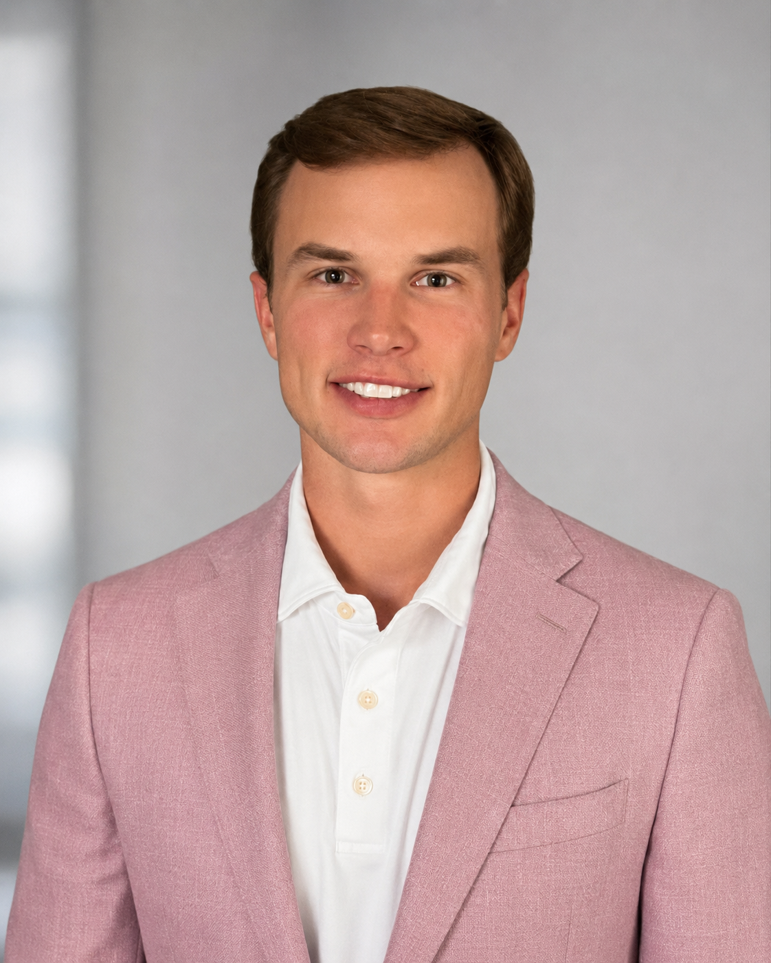 A young man with brown hair smiling, wearing a pink blazer and white shirt, standing in a professional setting.