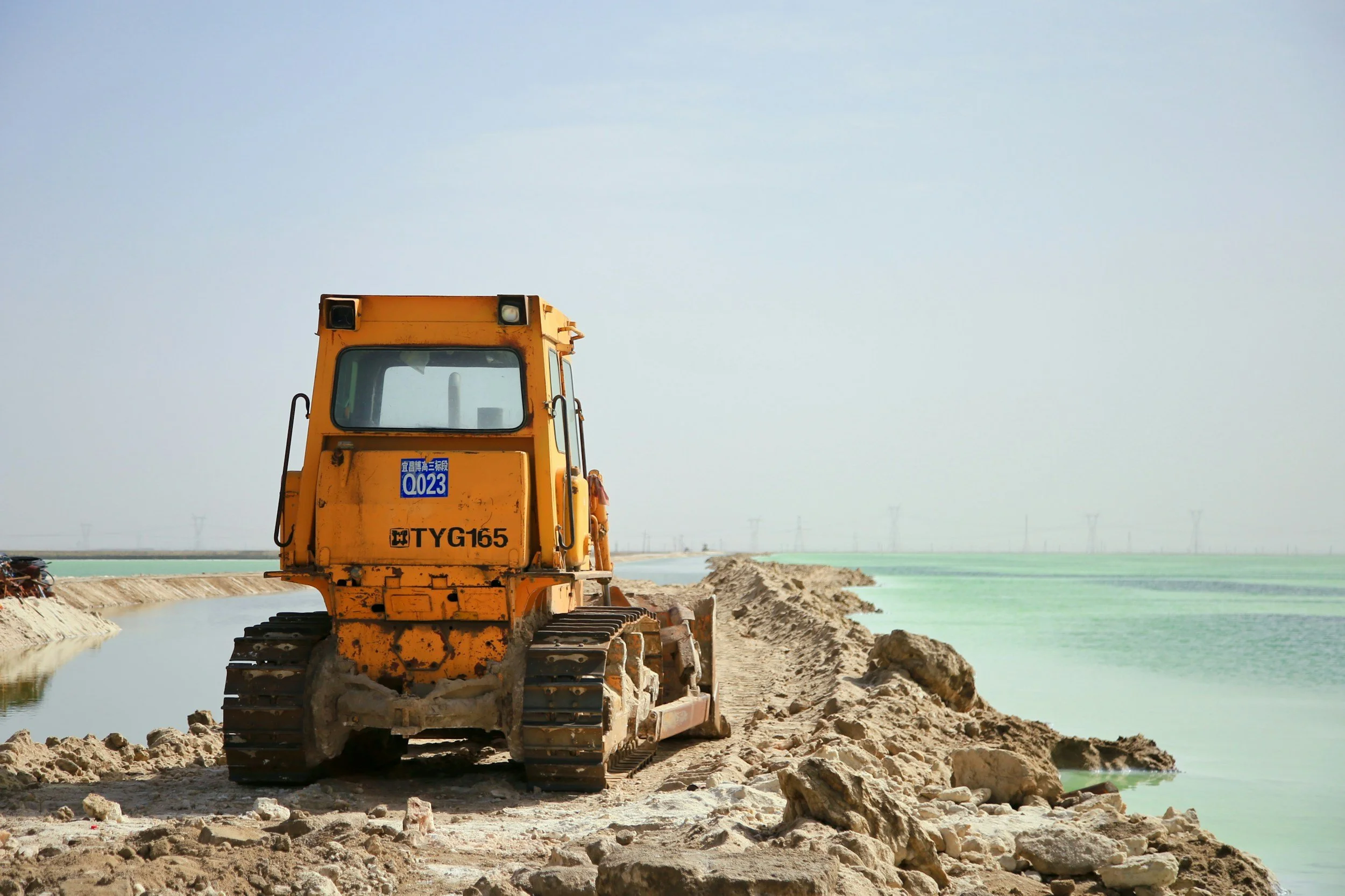 A yellow bulldozer on a construction site next to a body of water, with dirt and rocks on the ground and mountains in the background.