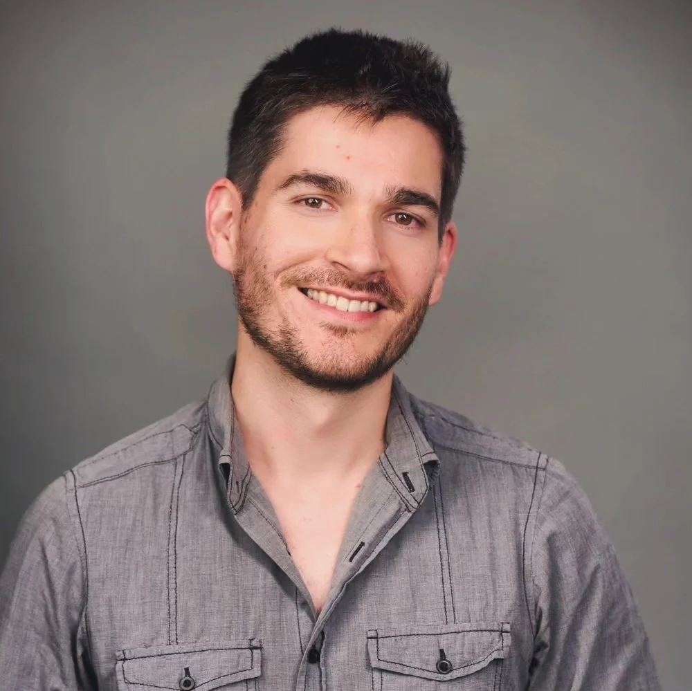 Murray, a young man with short dark hair and a beard, is smiling at the camera against a gray background. He is wearing a gray button-up shirt.