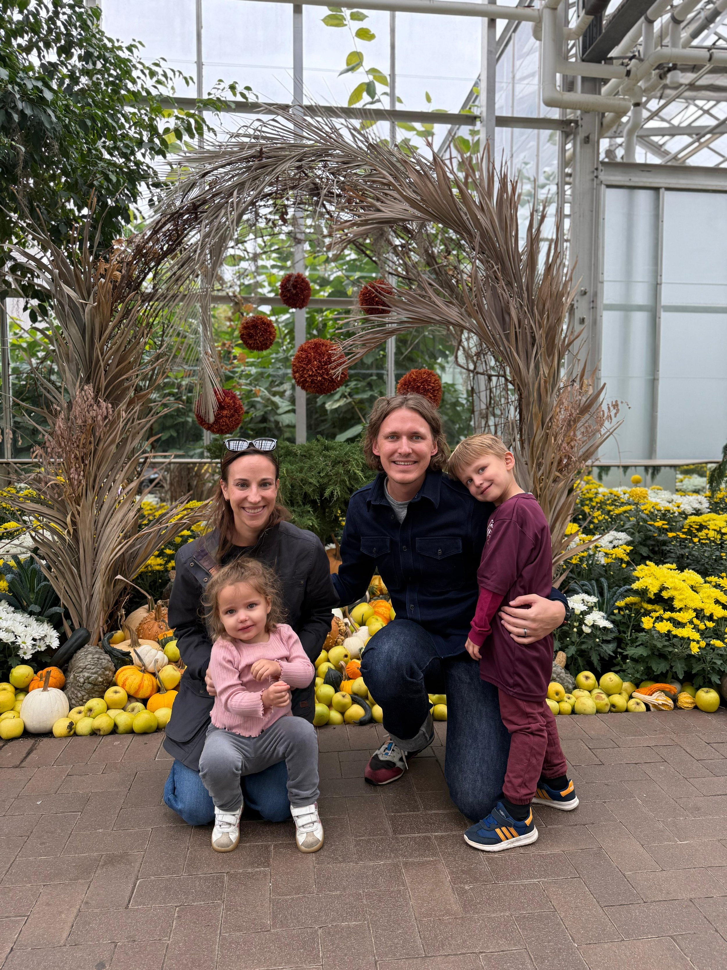 A photo of East Grand Rapids school board candidate Paul Bratt and his family: his wife, son, and daughter. They are smiling in front of an autumnal display at the Frederik Meijer Gardens in Grand Rapids, Michigan.
