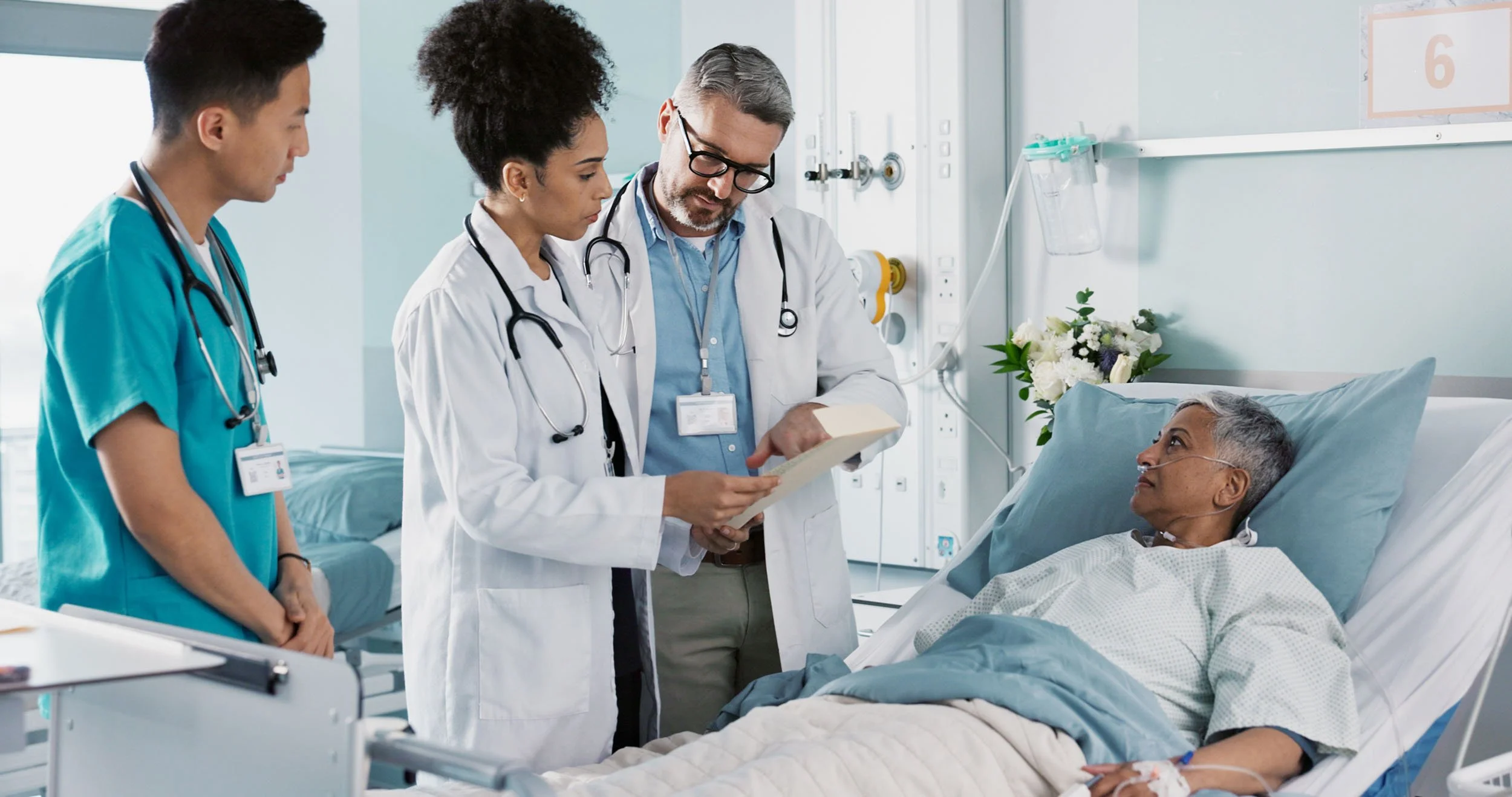 Medical team discussing a patient's treatment at the bedside in a modern hospital room, with two doctors and a nurse reviewing notes while speaking with the patient.