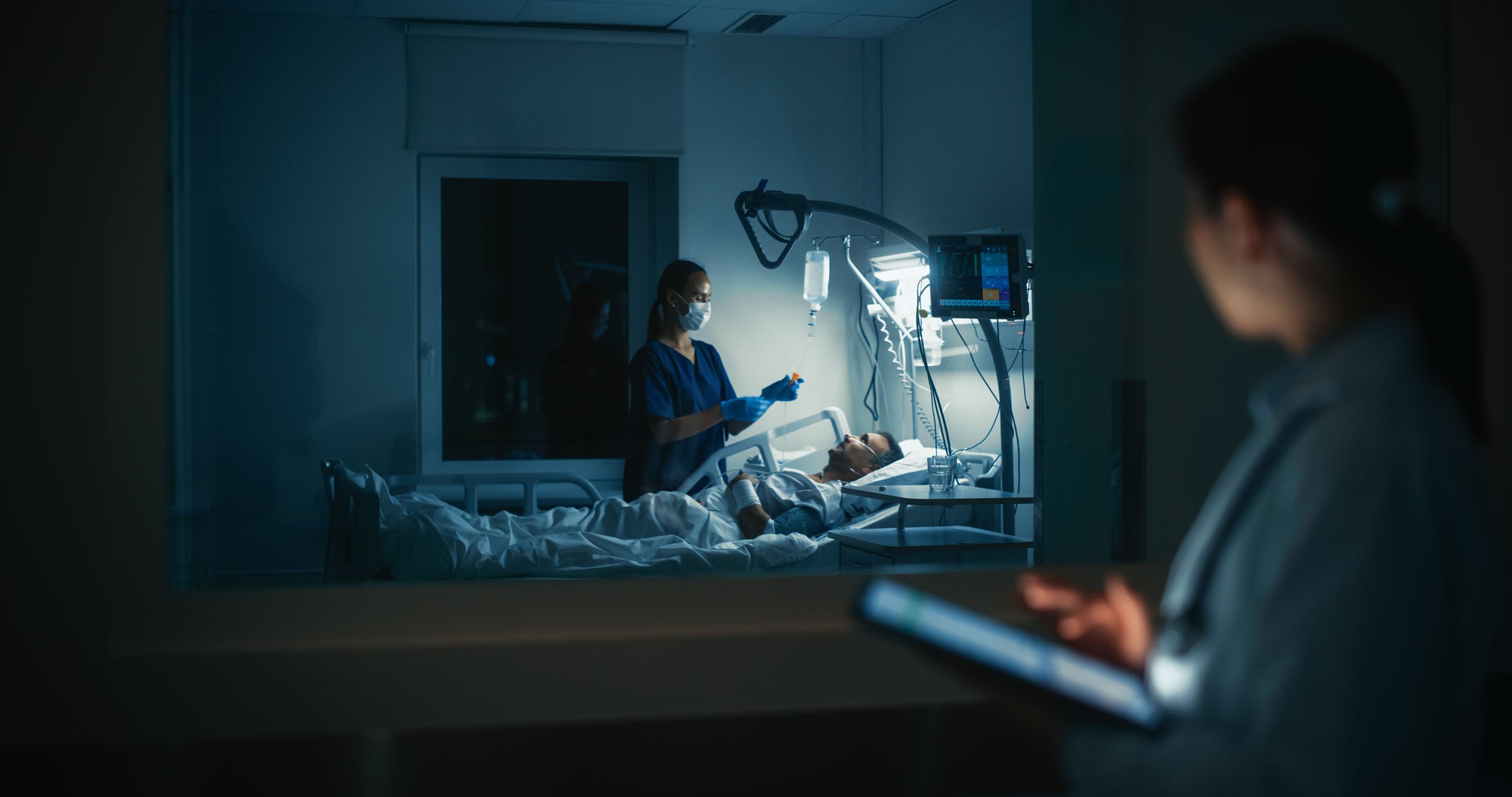 Nurse monitoring a patient receiving overnight care in a hospital room, viewed through an observation window, with medical equipment and vital signs monitor illuminated beside the bed.