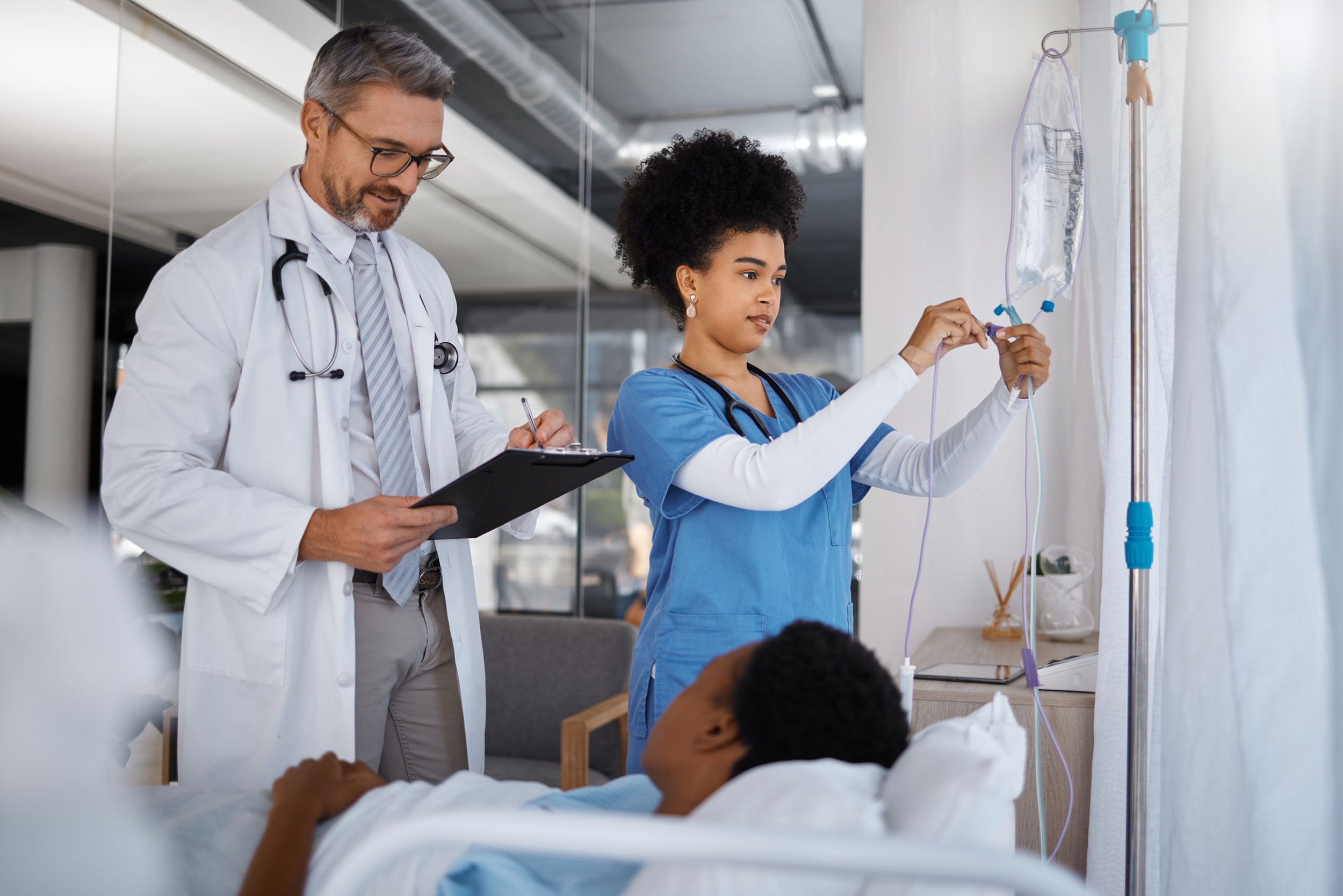 Doctor and nurse providing bedside care in a hospital room, with the nurse adjusting an IV drip while the doctor records patient information on a clipboard.