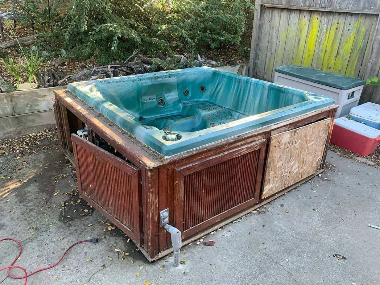 An outdoor hot tub with a blue interior and a wooden exterior, situated on a concrete patio near a fenced yard with plants and trees.