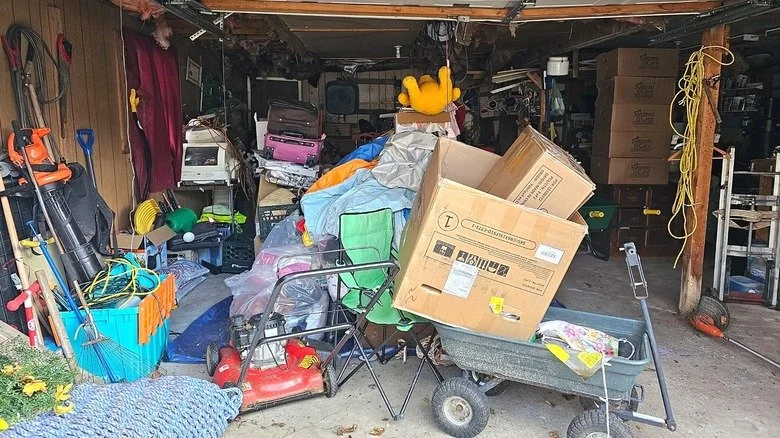A cluttered garage filled with various tools, boxes, and household items, including a lawn mower, plastic bins, a wheelbarrow, and scattered miscellaneous objects.