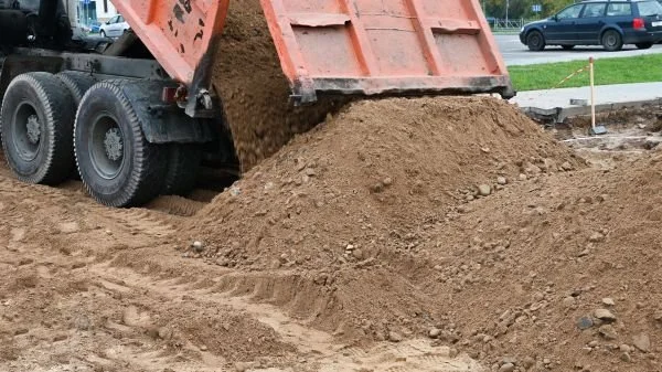 A dump truck unloading dirt onto a construction site with cars and a road in the background.