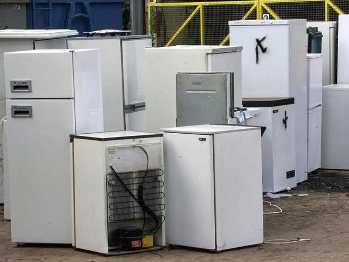 Various white refrigerators and freezers placed outdoors on a dirt surface near a yellow fence.
