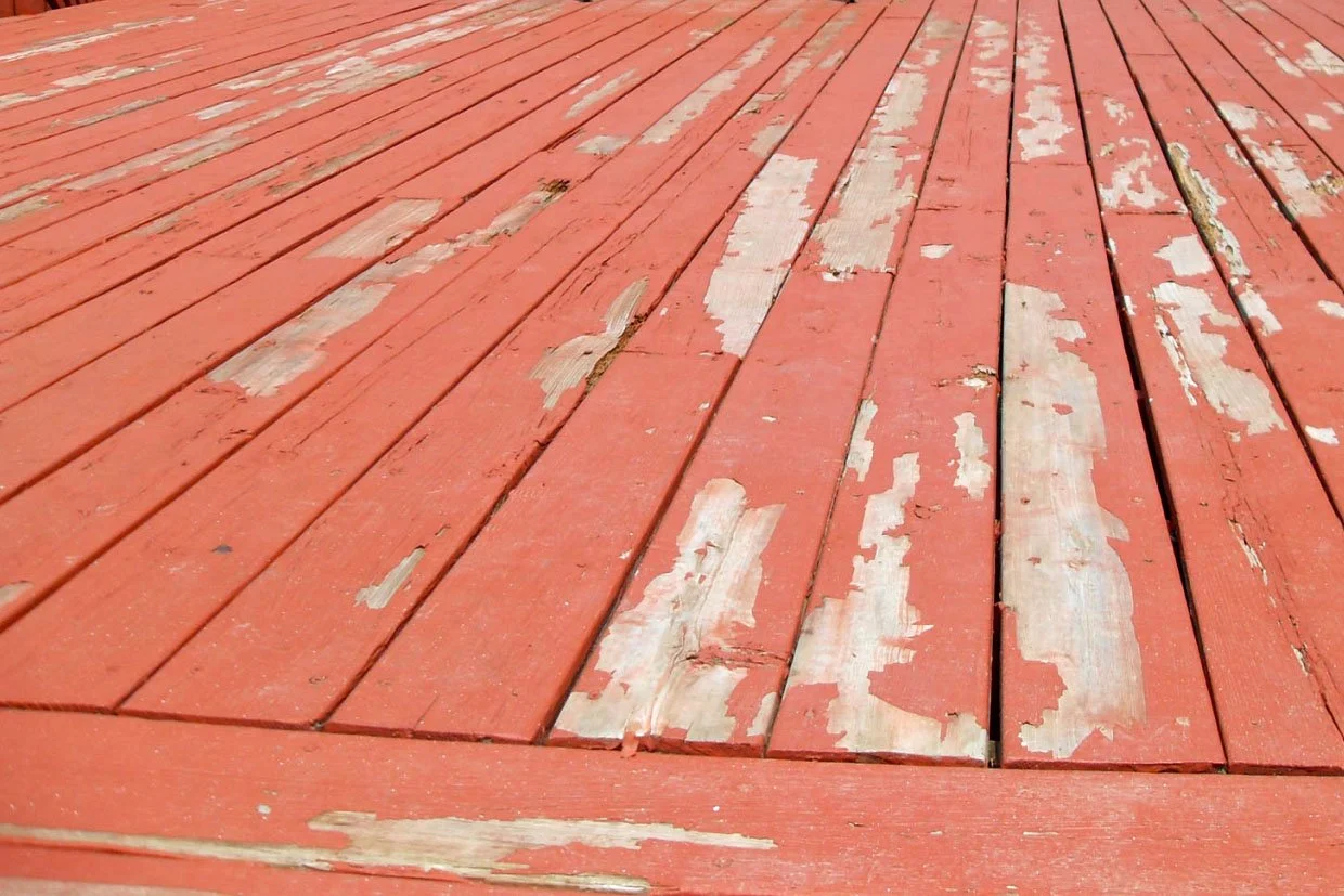 Close-up of a weathered, red-painted wooden deck with peeling paint and some damaged boards.