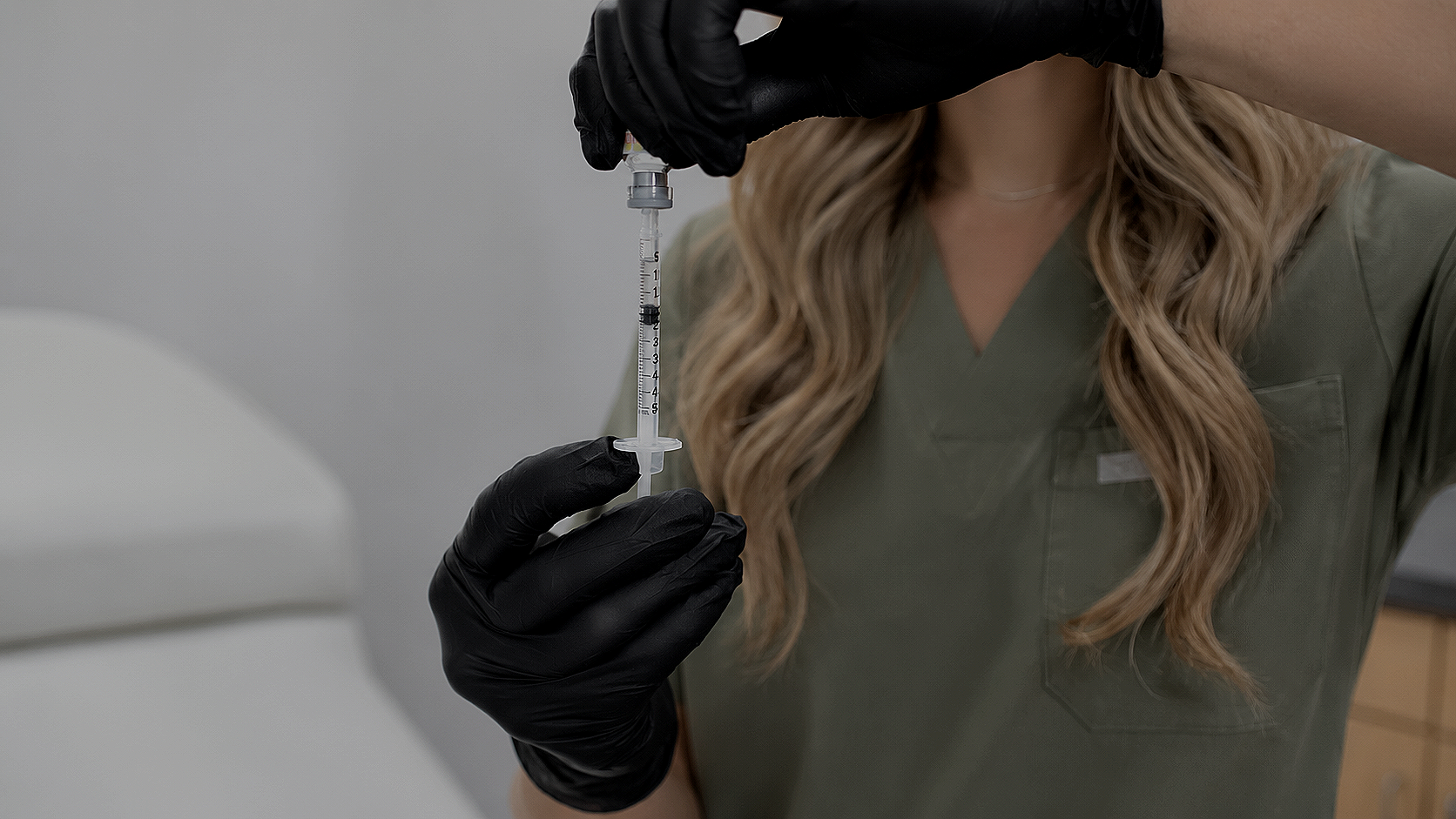 A healthcare worker in green scrubs and black gloves holding a syringe for injection.