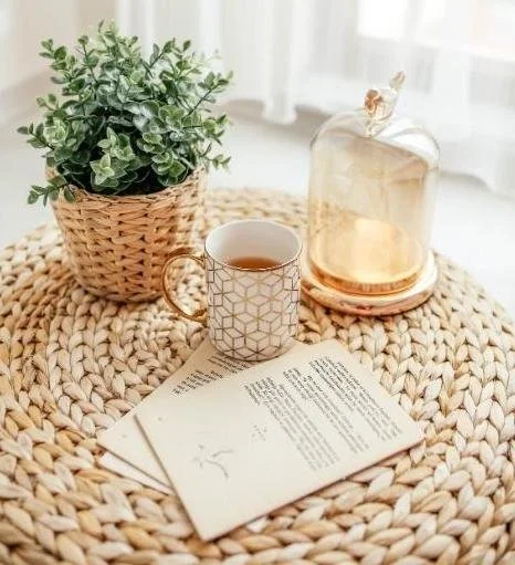 A cozy table scene with a potted green plant in a woven basket, a cup of tea on a book, and a glass teapot with a lid, all on a round woven placemat.