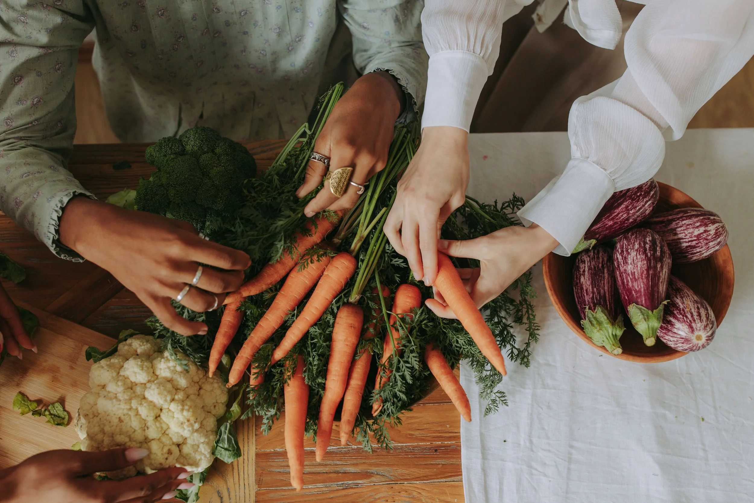 Hands of two people picking fresh carrots from a basket filled with carrots. There are also broccoli, cauliflower, and eggplants on the table. A whole-food approach to holistic eating