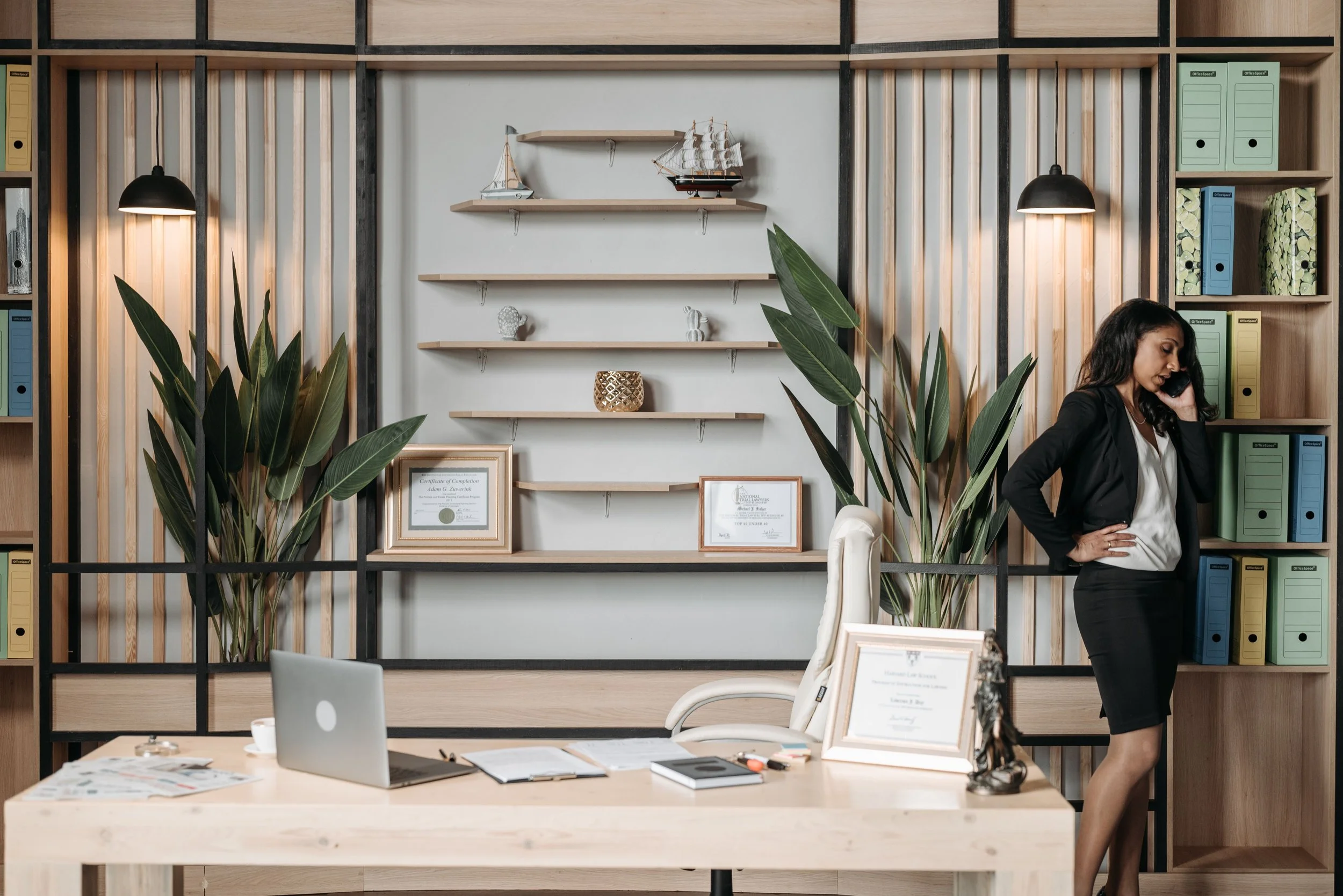 A registered dietitian in business attire talking on the phone in an office with a modern wooden and black metal shelf, plants, books, and decorative items in the background.