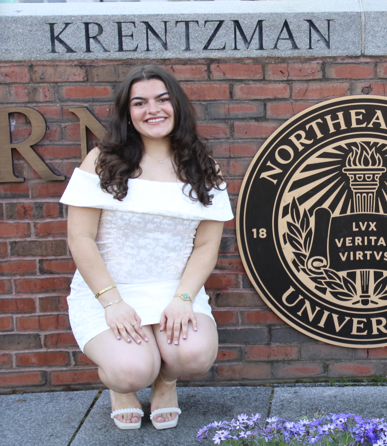 A young woman with long dark curly hair and light skin posing outside in front of a brick wall with university signage. She is wearing a white off-the-shoulder dress and white sandals adorned with pearls, with jewelry including bracelets, a watch, and a necklace, smiling at the camera.