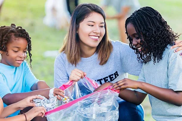 Three girls, two teenagers and a young girl, sitting outside on grass, smiling, and holding a clear plastic bag with aluminum foil inside.