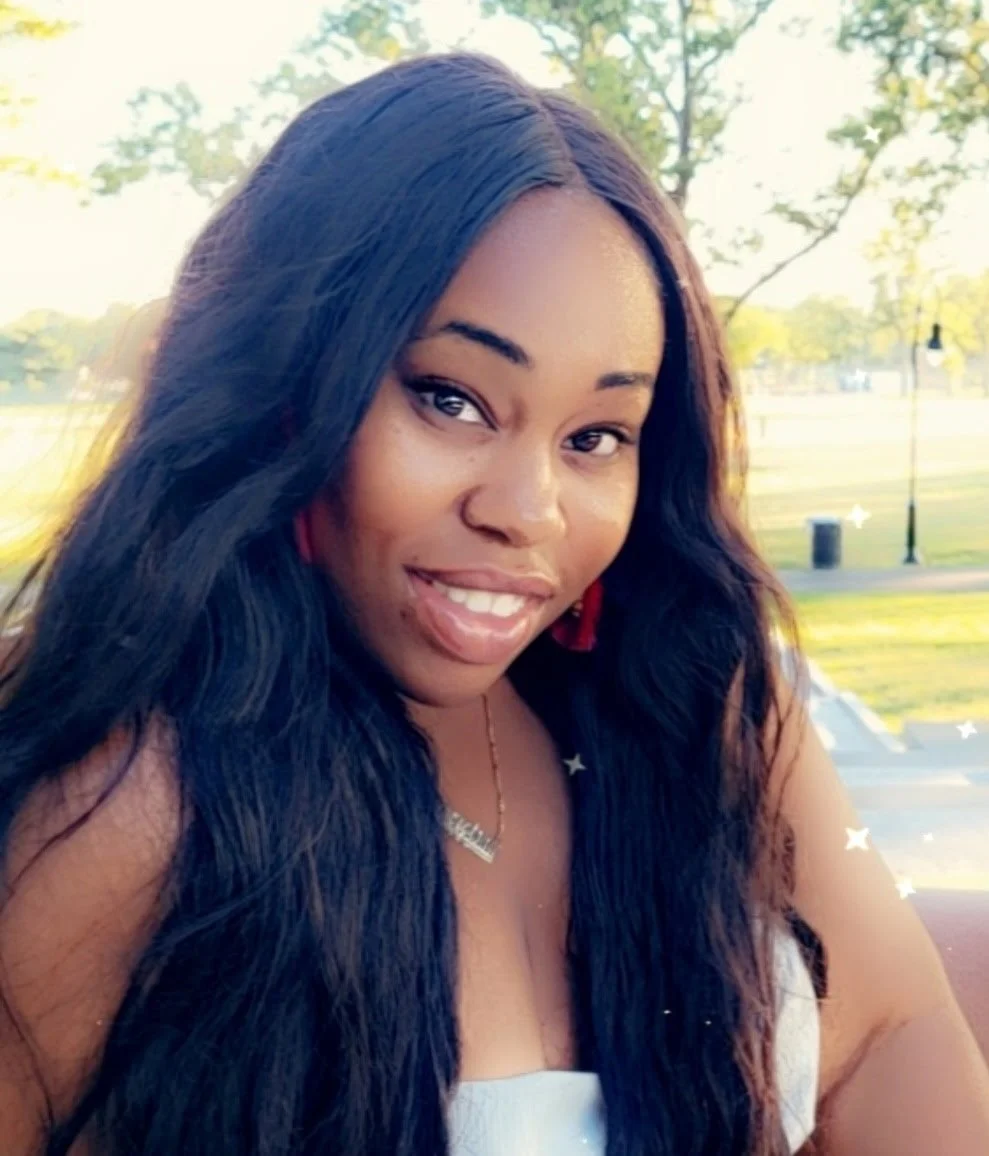Young woman with long black hair smiling outdoors during daytime, with trees and park in background.